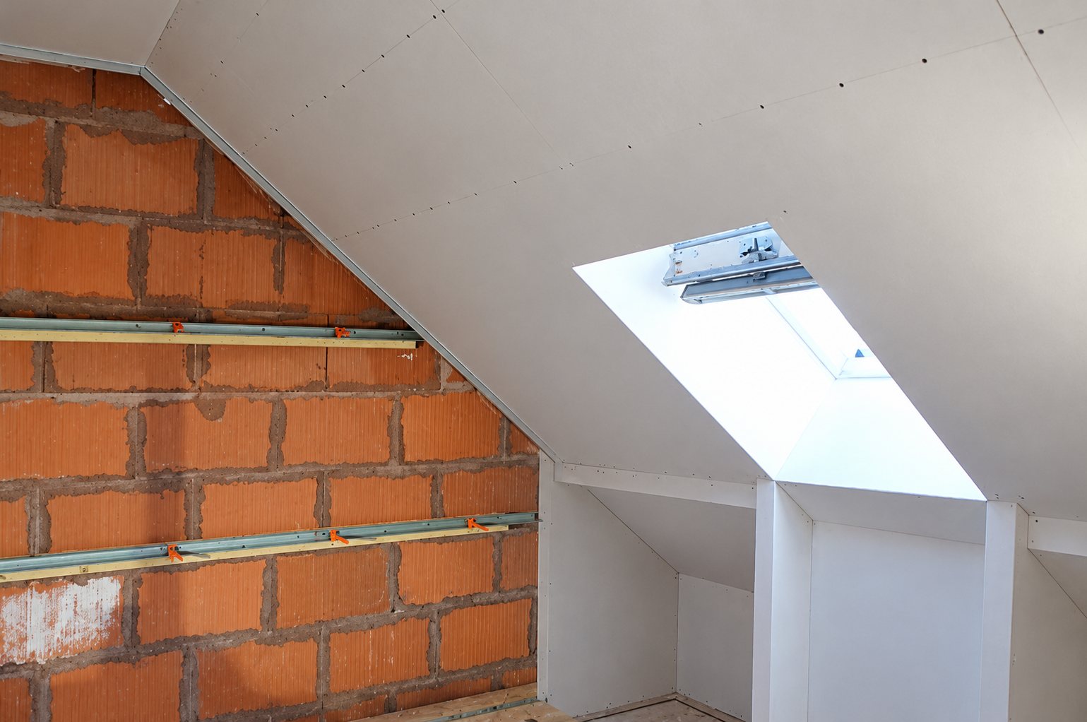 Combles en cours de rénovation avec plaques de plâtre posées sous la pente du toit, une fenêtre de toit et un mur de briques rouges avec rails métalliques.