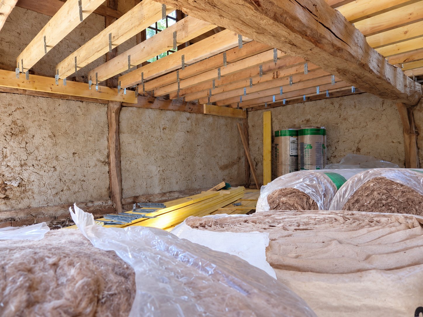 Sous un plancher en rénovation, des solives en bois neuves surplombent des murs en pierre et des rouleaux d'isolant emballés dans du plastique.
