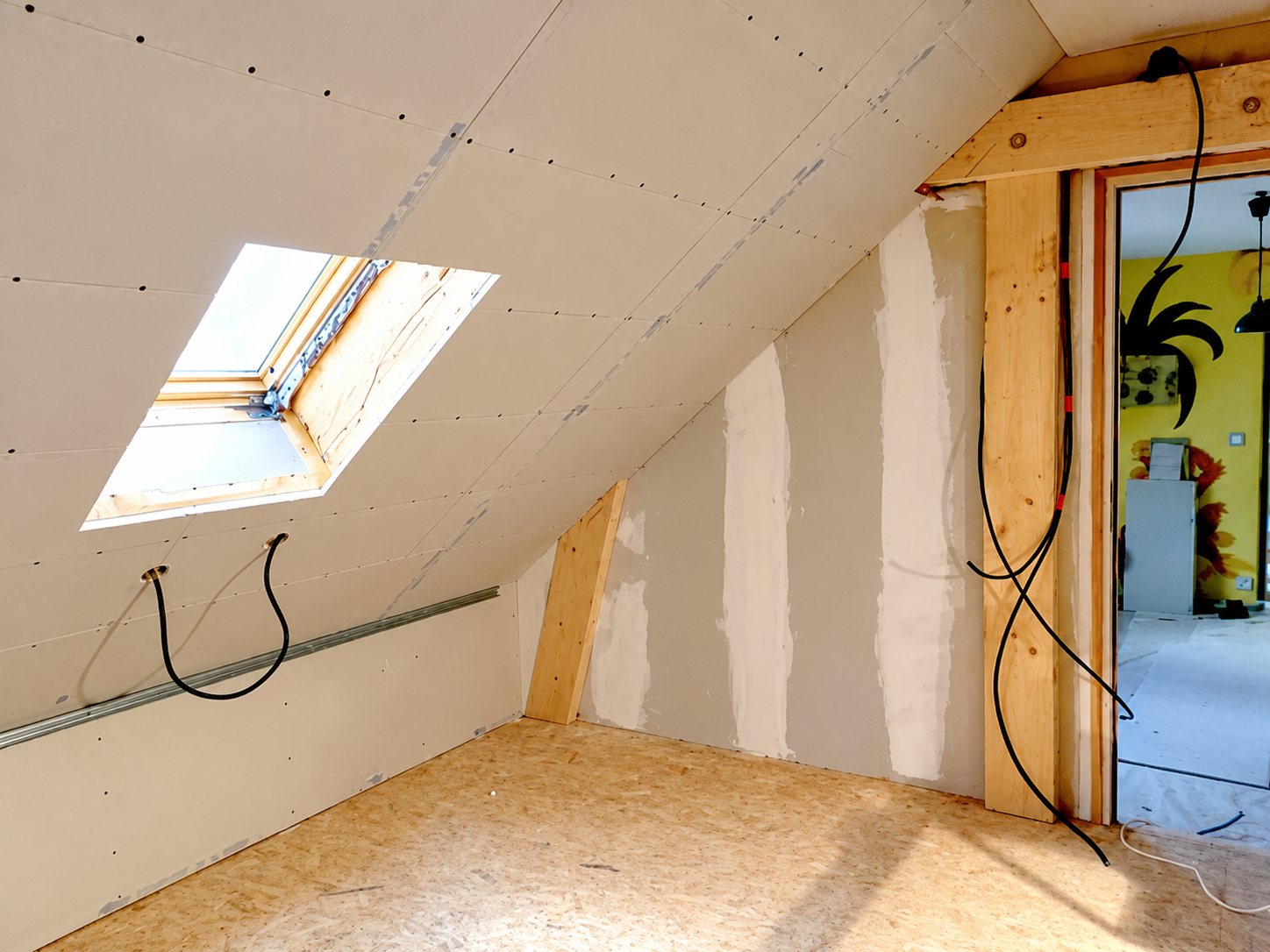 Pièce sous les combles en travaux, avec murs en plaques de plâtre, sol en panneaux OSB, fenêtre de toit et câbles électriques apparents.
