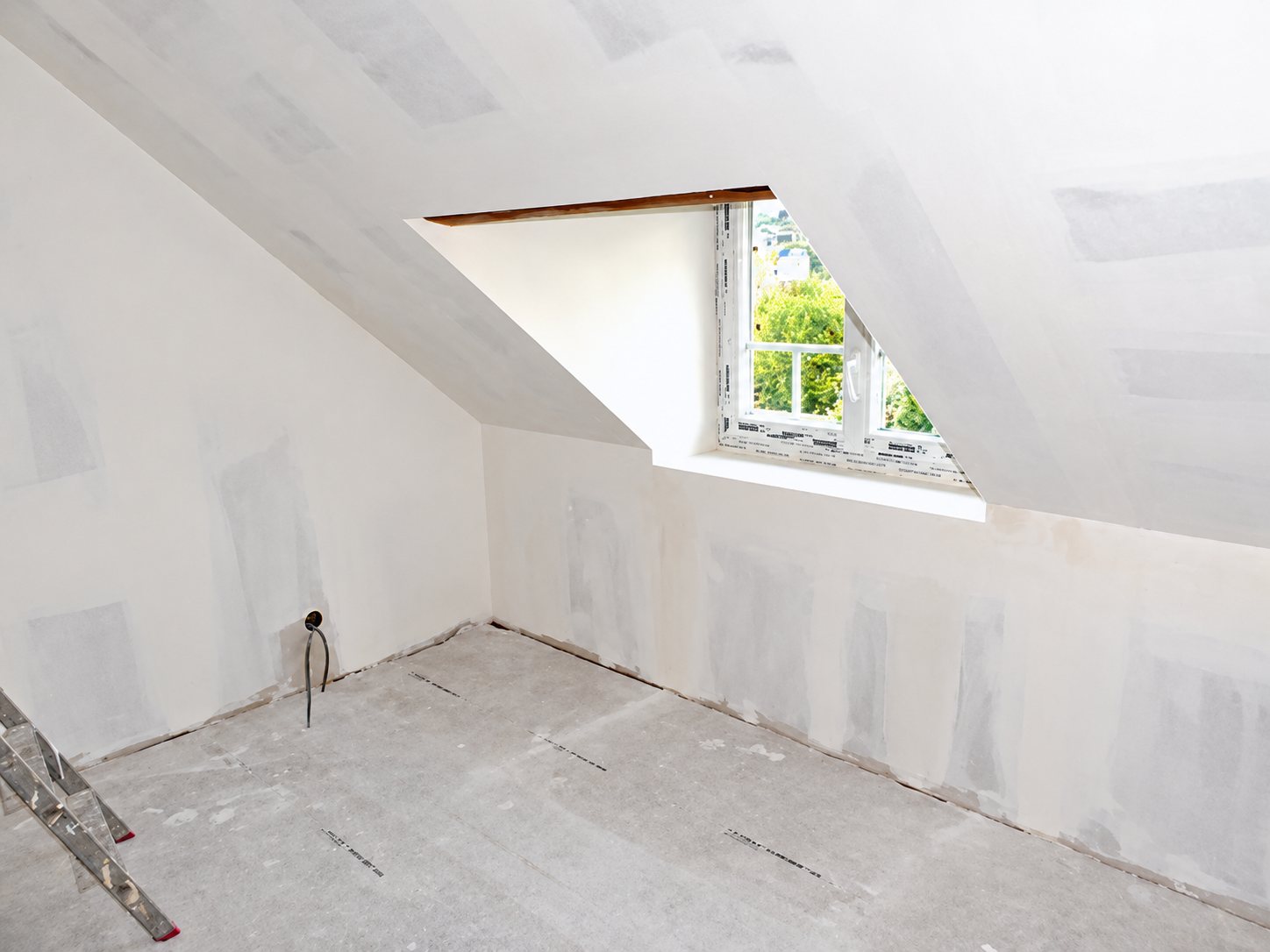 Pièce sous les combles en travaux, aux murs et plafond en plâtre enduit, avec une lucarne donnant sur la verdure et une échelle au premier plan.