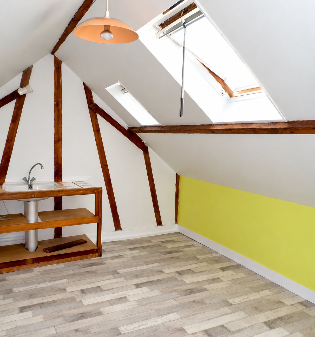 Pièce sous les combles rénovée avec poutres en bois apparentes, deux fenêtres de toit, un mur jaune, un sol imitation parquet clair et un lavabo sur un meuble en bois.
