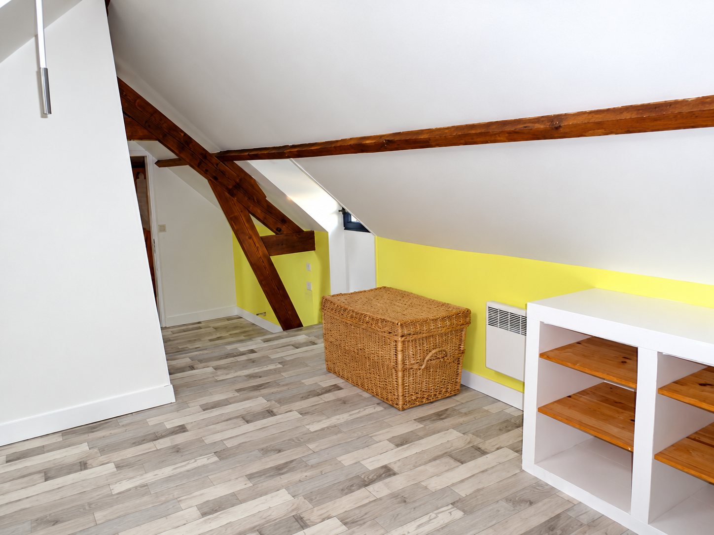 Pièce sous les combles rénovée avec poutres en bois apparentes, mur jaune, sol stratifié gris clair, malle en osier et étagère blanche.