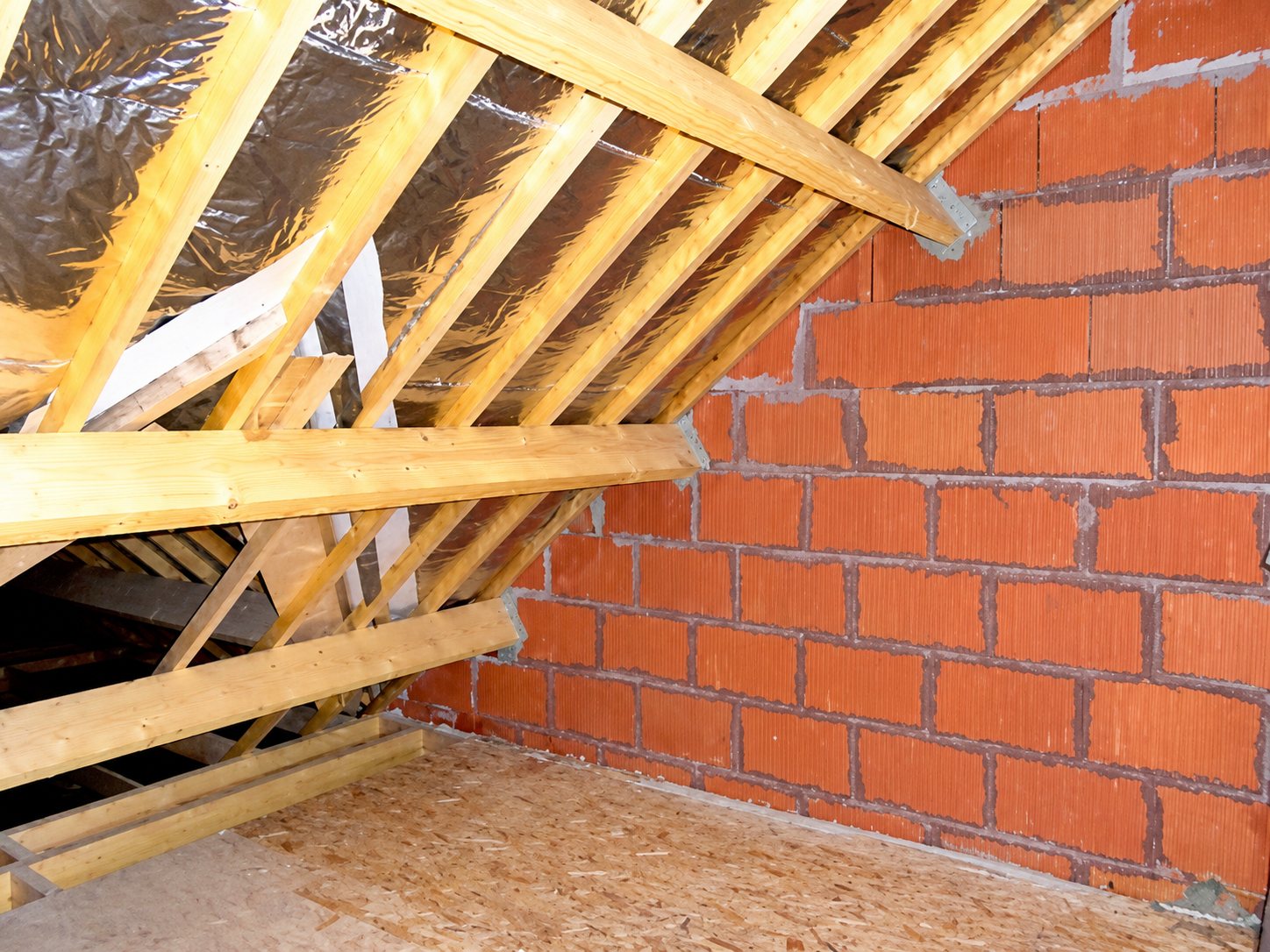 Intérieur de combles en chantier avec une charpente en bois, une isolation réfléchissante sous la toiture et un mur de briques rouges.