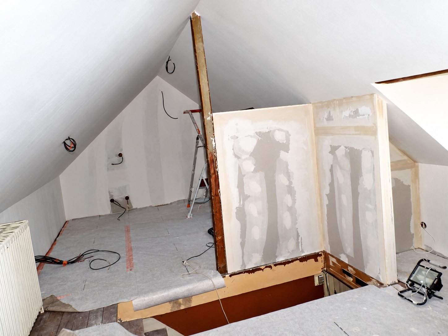 Des combles aménagés en chantier, avec des cloisons en plaques de plâtre jointoyées, un projecteur de chantier, des câbles au sol et un escabeau.