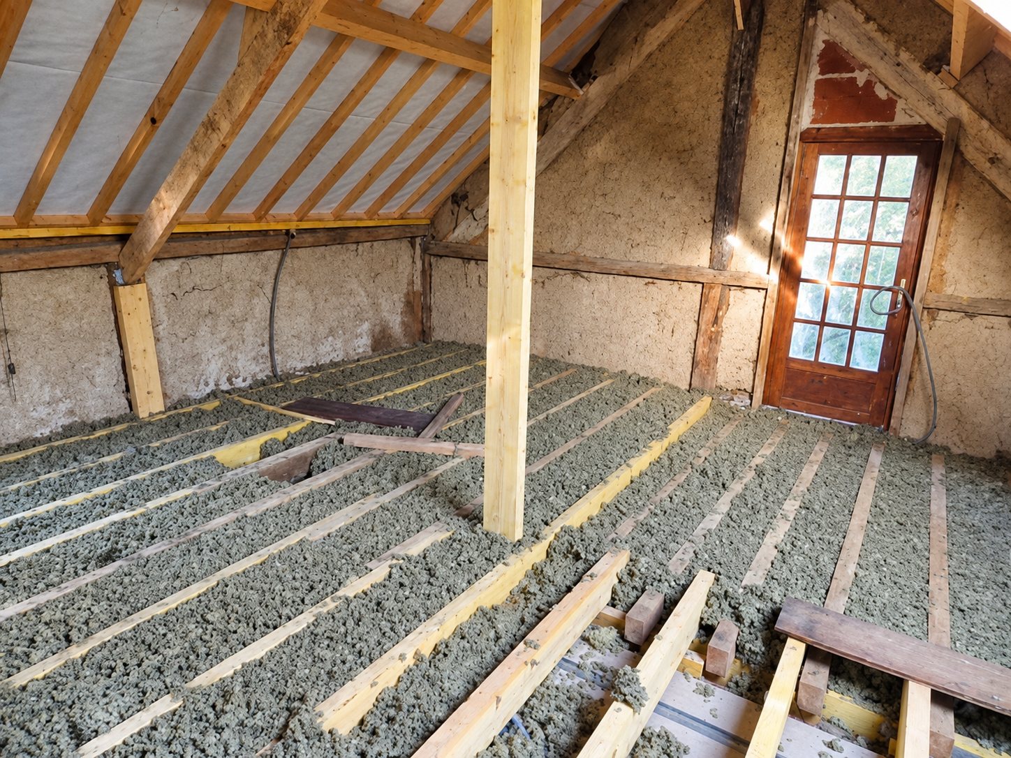 Vue intérieure de combles dont le sol est recouvert d'isolant en ouate de cellulose entre des solives en bois, avec charpente apparente et une porte vitrée au fond.