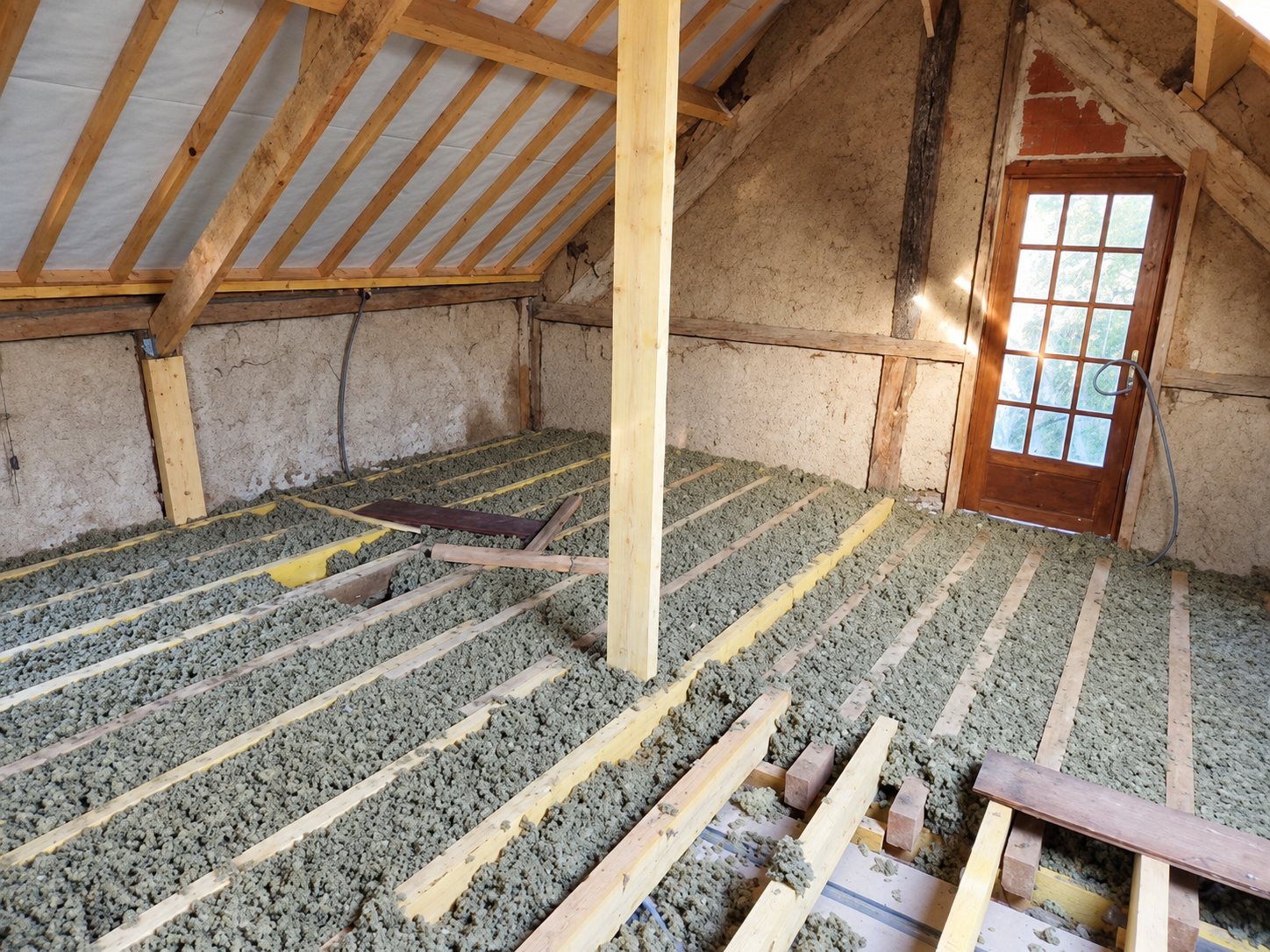 Intérieur de combles en cours d'isolation, avec de la ouate de cellulose soufflée entre les solives en bois, une charpente apparente et une porte vitrée au fond.