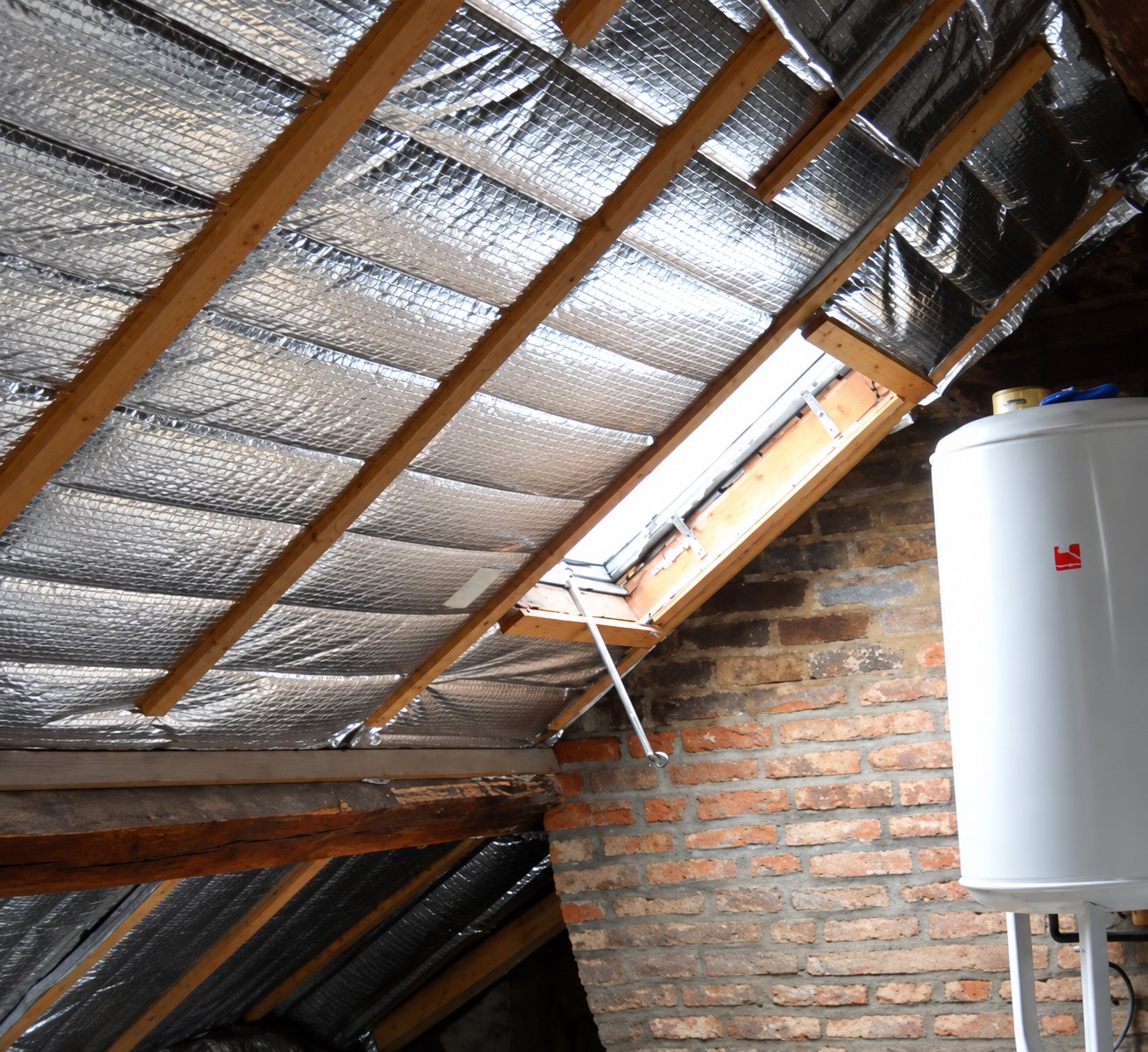 Sous la toiture, des chevrons en bois sont recouverts d'un isolant réflecteur argenté, à côté d'une fenêtre de toit, d'un mur en briques et d'un chauffe-eau blanc.