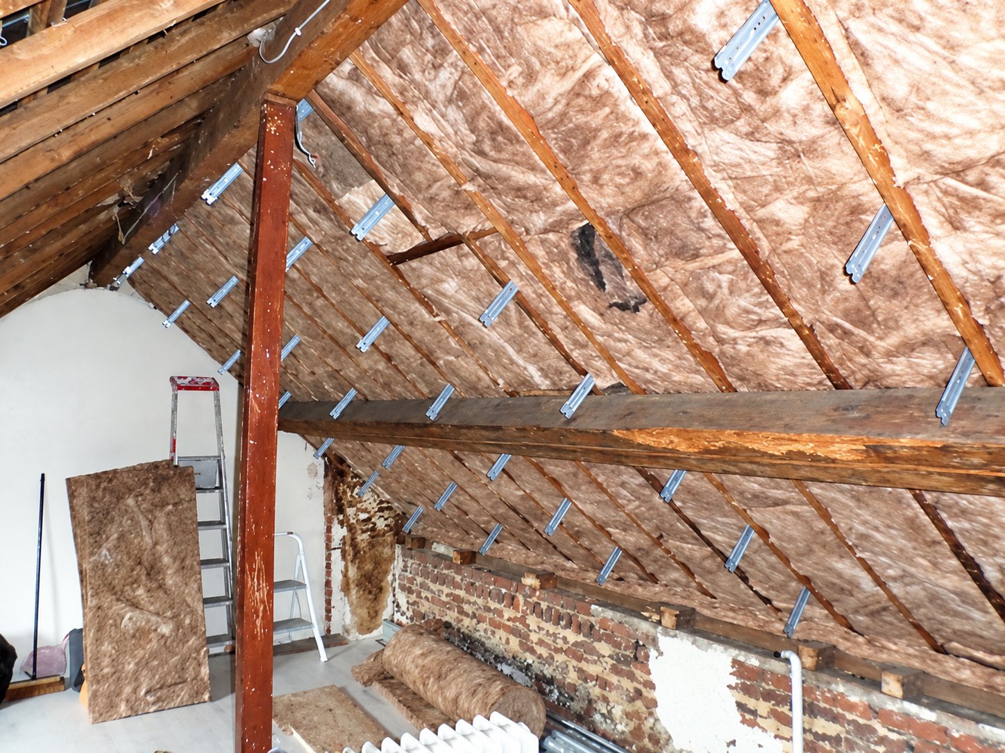 Vue de combles en cours de rénovation, avec une charpente en bois apparente, de la laine de verre posée entre les chevrons et des appuis métalliques de fixation.