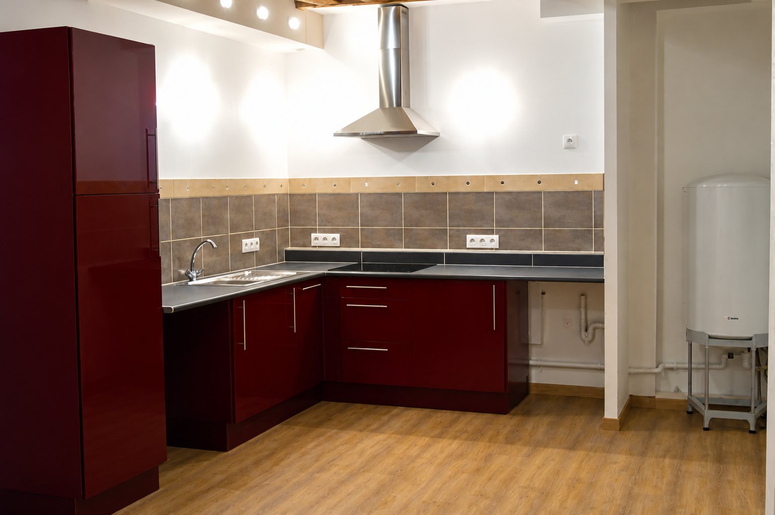 Cuisine récemment aménagée avec meubles bordeaux, plan de travail gris, crédence en carrelage et hotte en inox, à côté d'un chauffe-eau.