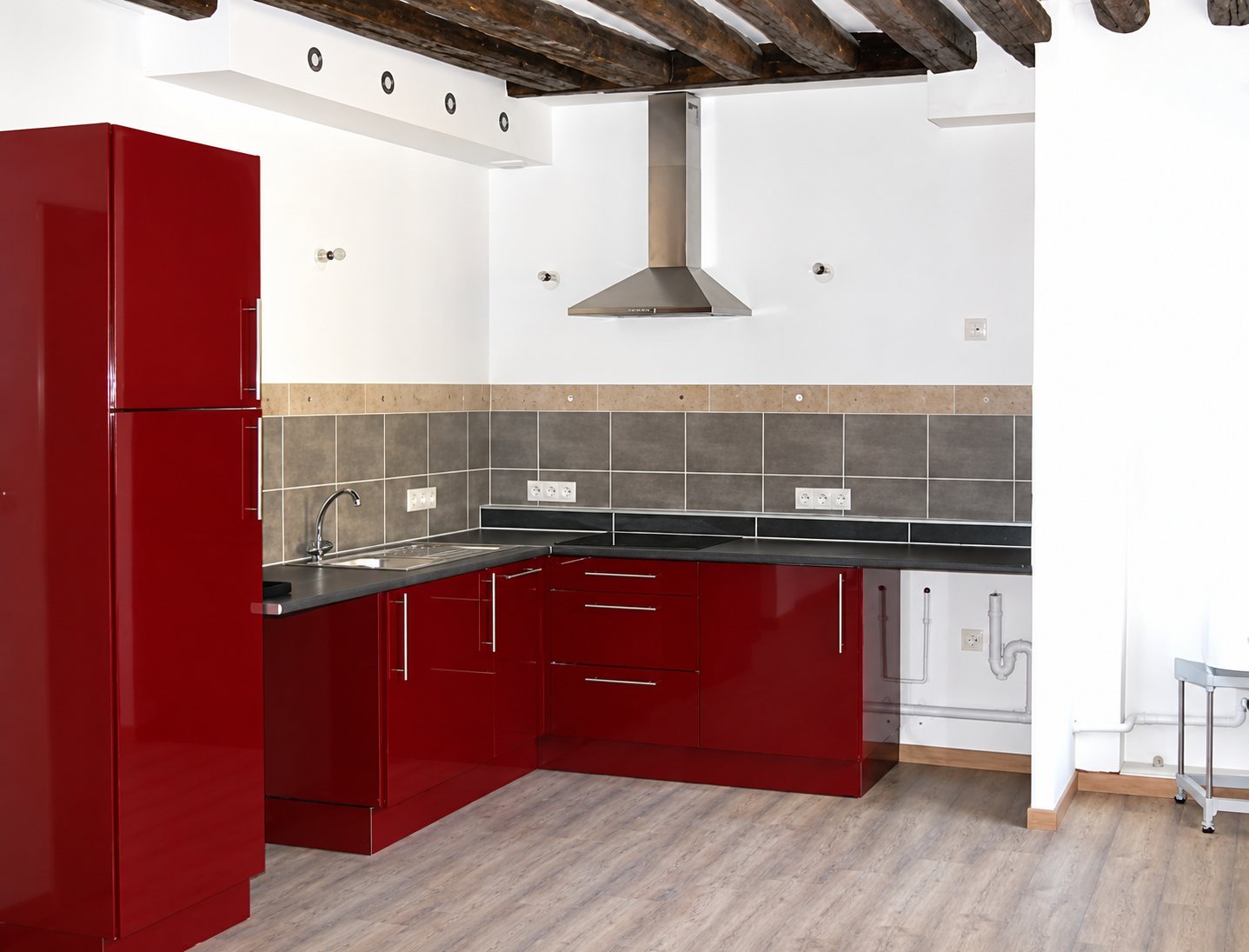 Cuisine moderne en angle aux meubles rouge laqué, avec crédence grise, hotte inox et plafond à poutres apparentes.