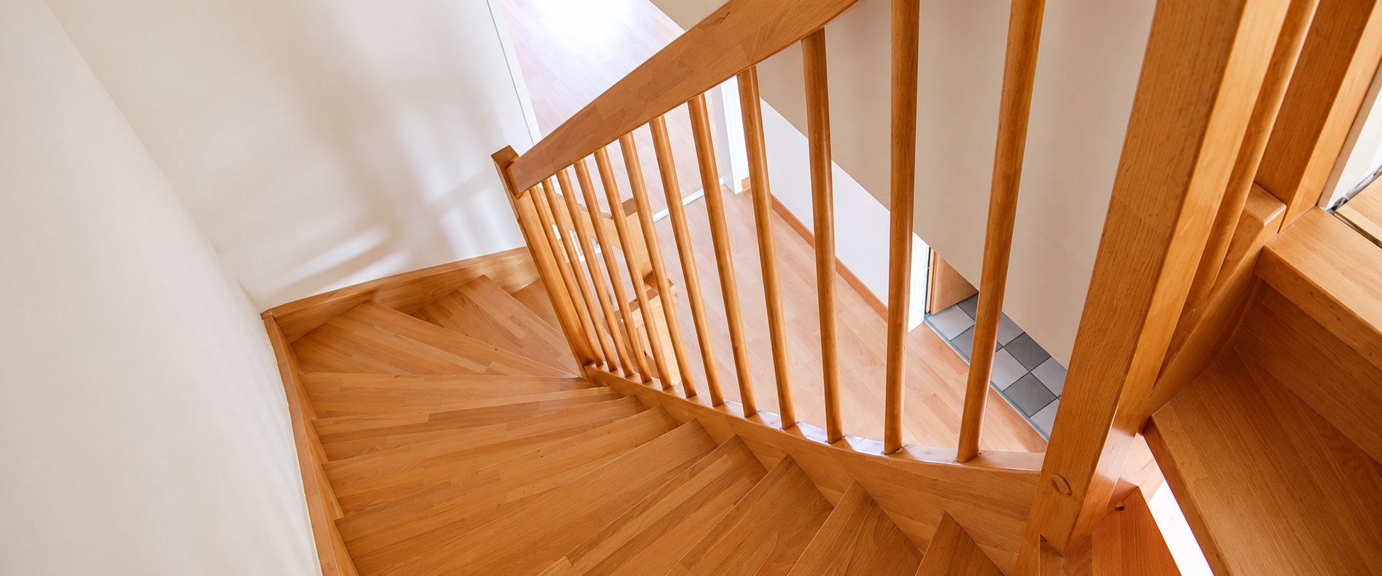 Vue plongeante sur un escalier tournant en bois clair avec rampe à barreaux verticaux, dans un intérieur aux murs blancs.