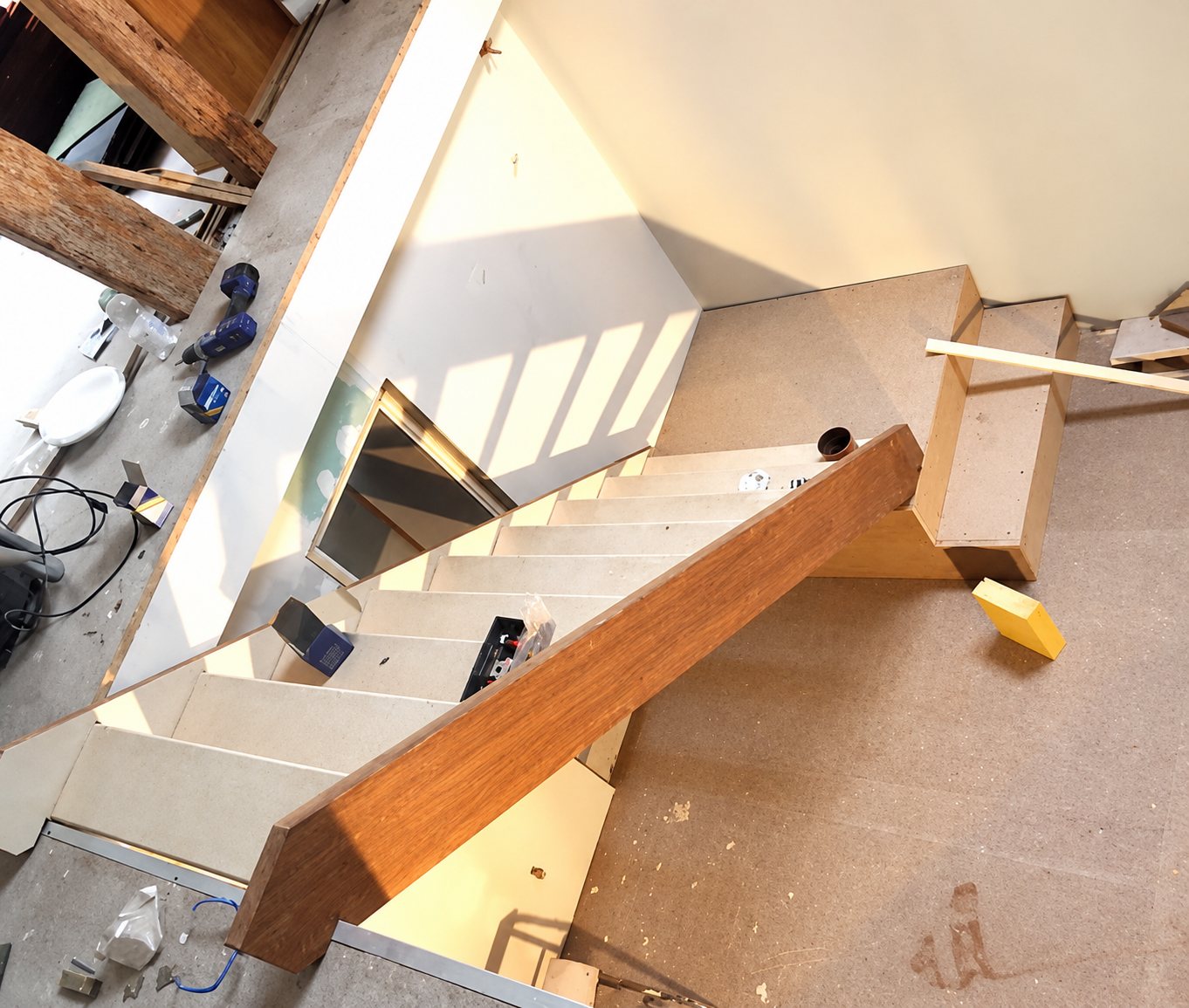 Vue plongeante sur un escalier en bois clair avec limon en bois foncé, en cours d'installation sur un chantier intérieur avec des outils posés autour.