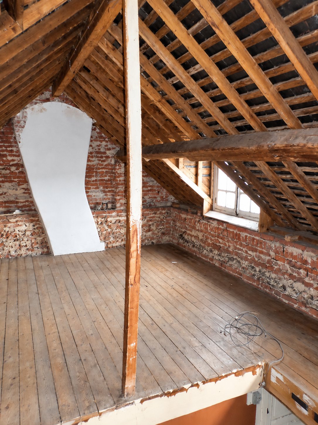 Vaste grenier aménagé sous une charpente en bois apparente, avec des murs en briques, un parquet ancien et une petite lucarne.