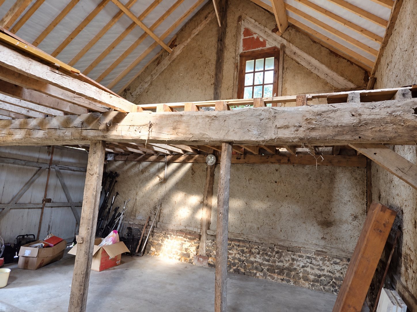 Intérieur d'une vieille grange en cours de rénovation, avec de grosses poutres en bois, une charpente neuve et des murs en pierre et en torchis.