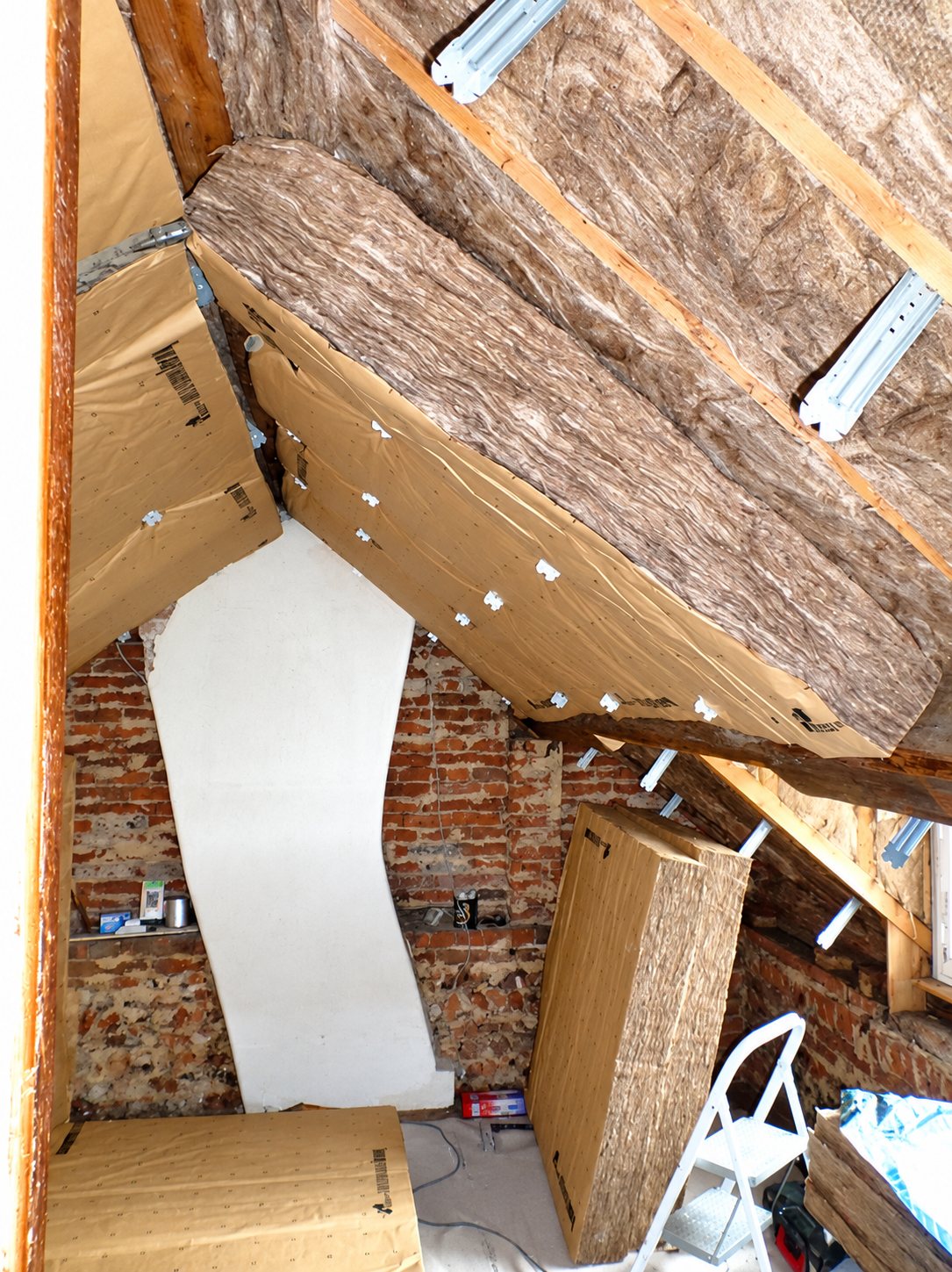 Des panneaux de laine isolante sont posés sous la charpente d'un comble en rénovation, avec un mur de briques apparentes et un escabeau au premier plan.