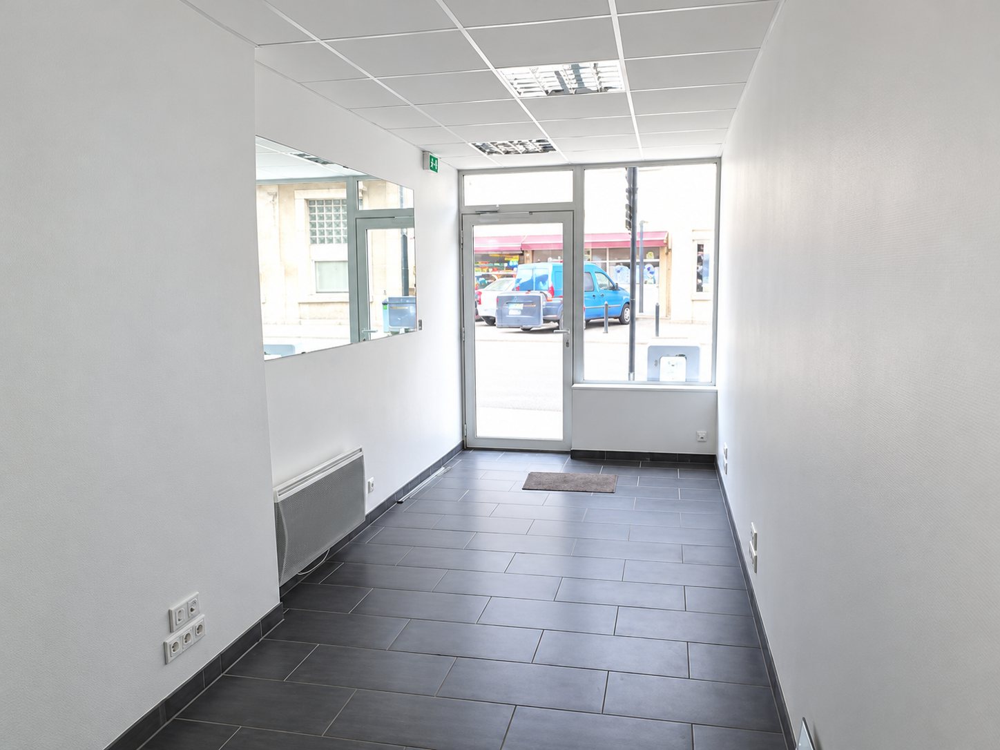 Couloir lumineux d'un local commercial vide, au sol carrelé gris et aux murs blancs, avec une porte vitrée donnant sur la rue.