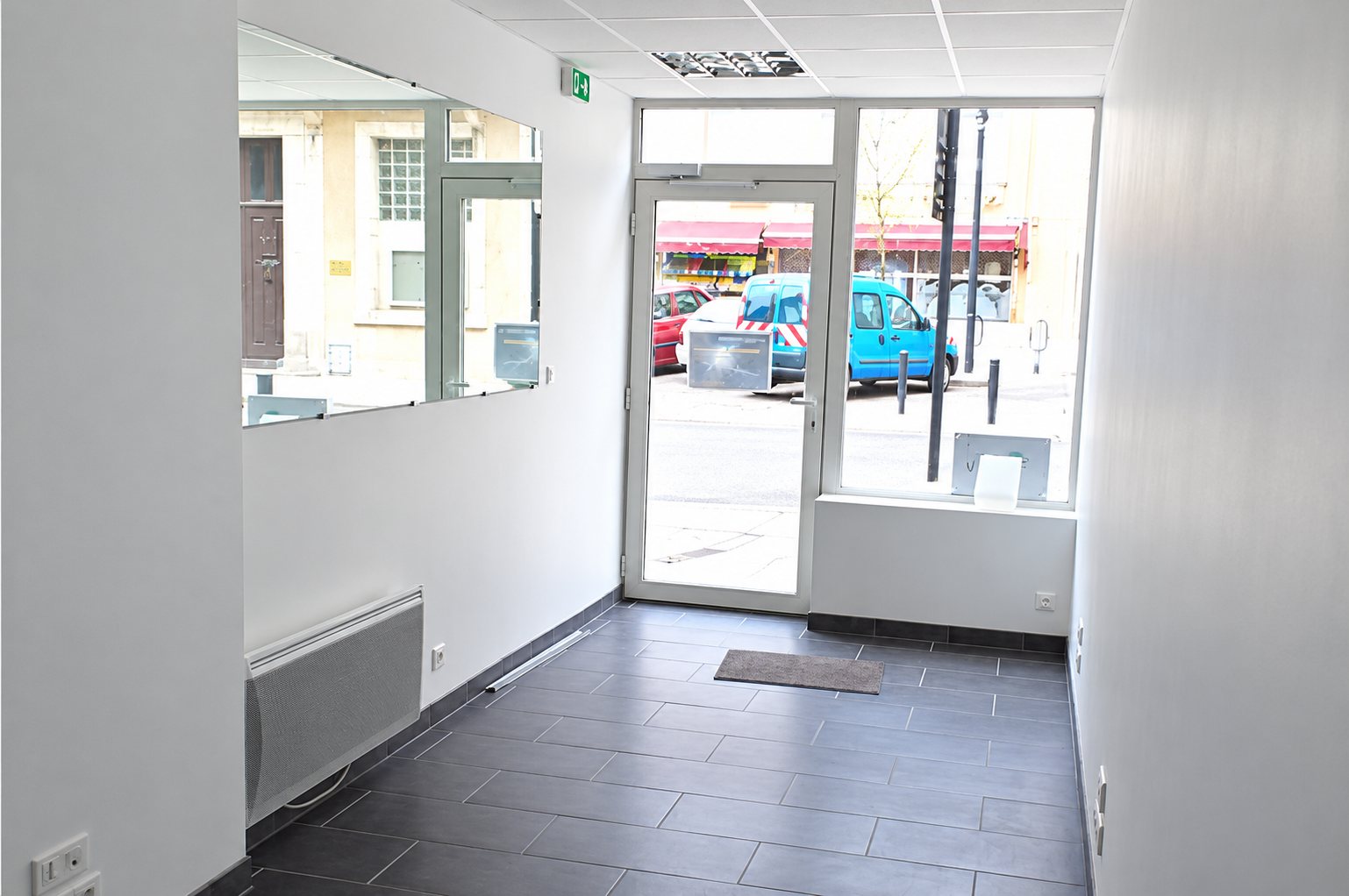 Intérieur d'un petit local commercial vide aux murs blancs, avec un carrelage gris, un radiateur, un grand miroir et une porte vitrée donnant sur la rue.
