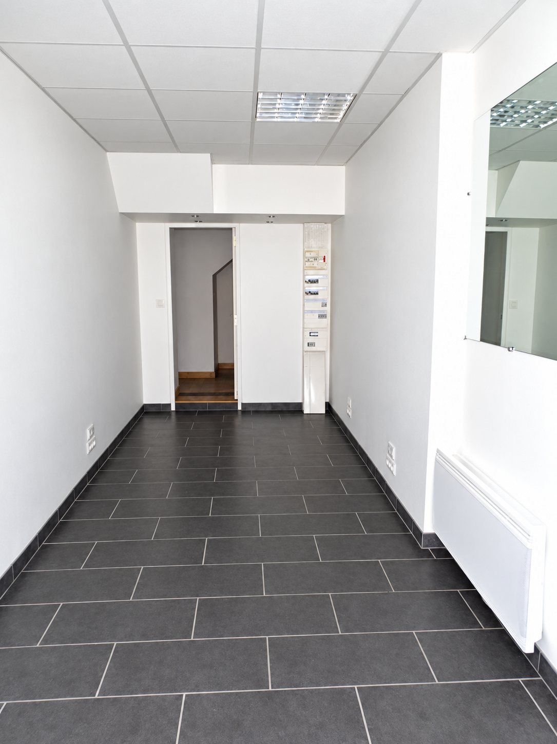Intérieur d'un local vide aux murs blancs, avec un sol en carrelage gris foncé, un miroir à droite et une porte ouverte au fond sur un escalier.