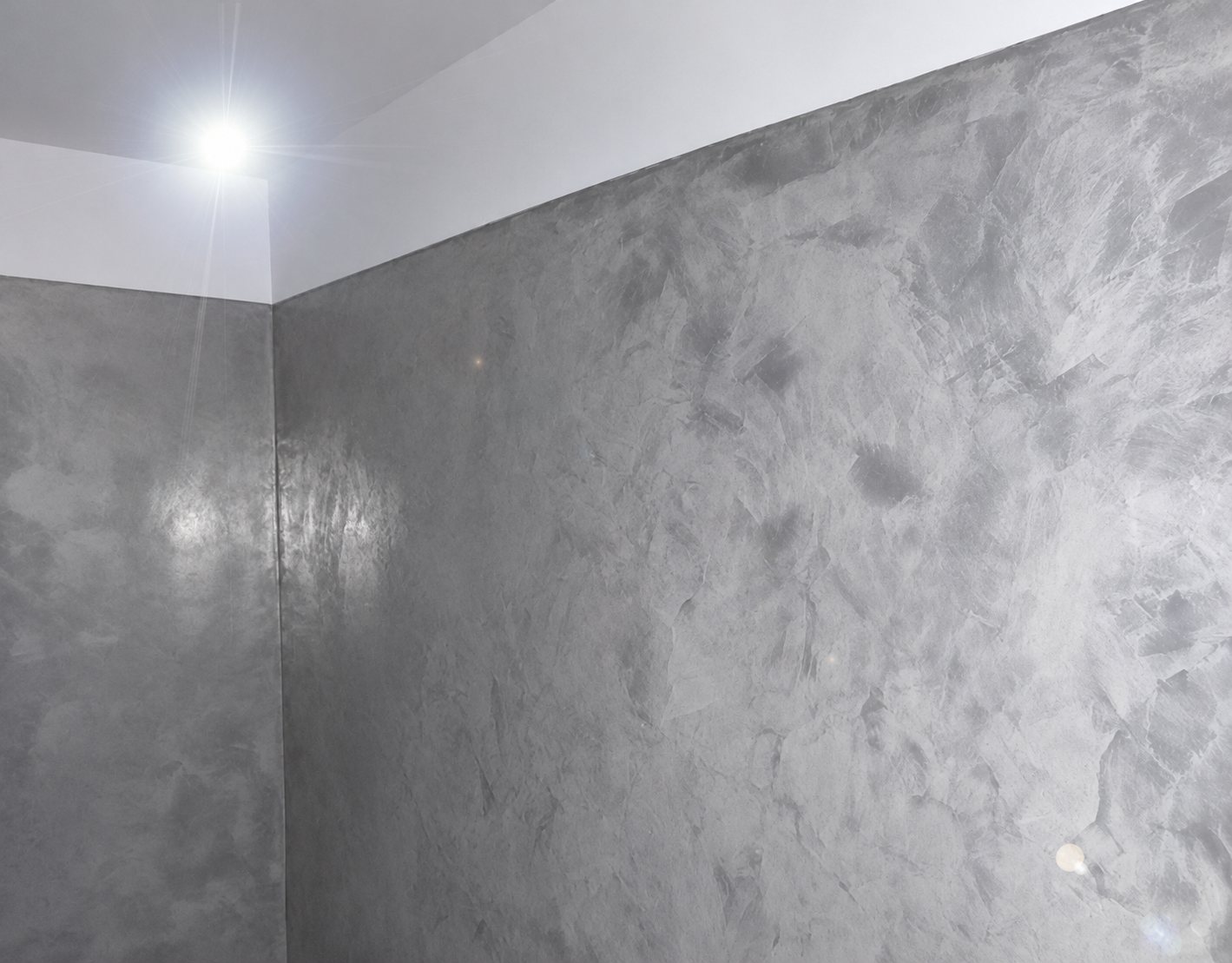Angle d'une pièce dont les murs sont recouverts d'un enduit gris nuancé de type béton ciré, éclairé par une lampe au plafond.