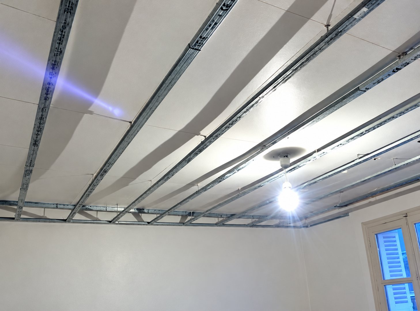 Rails métalliques d'un faux plafond en cours de pose dans une pièce aux murs blancs, avec une ampoule allumée et une fenêtre à volets bleus.