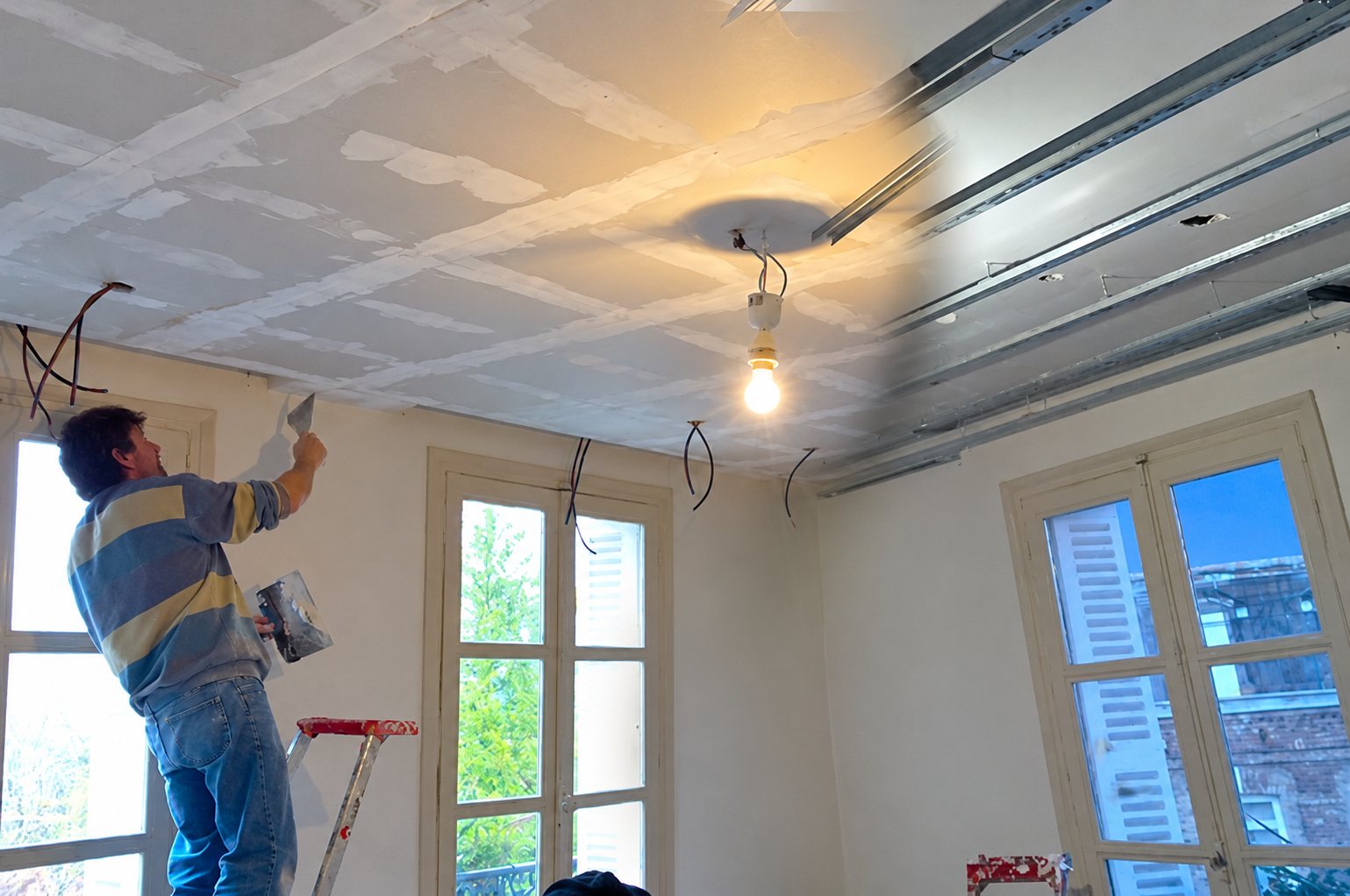 Un plâtrier enduit les joints d'un plafond en plaques de plâtre dans une pièce en rénovation, avec une ampoule nue et de grandes fenêtres.