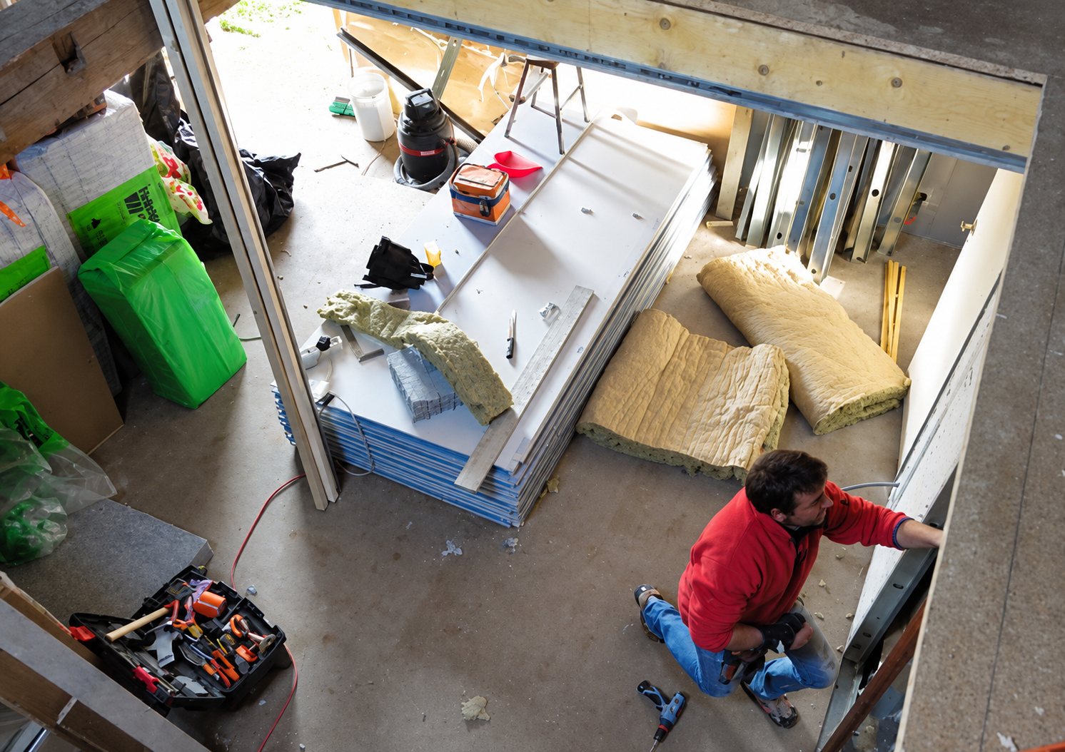 Vue en plongée d'un ouvrier en pull rouge agenouillé qui fixe une cloison dans un chantier intérieur, à côté d'une pile de plaques de plâtre et de rouleaux d'isolant.