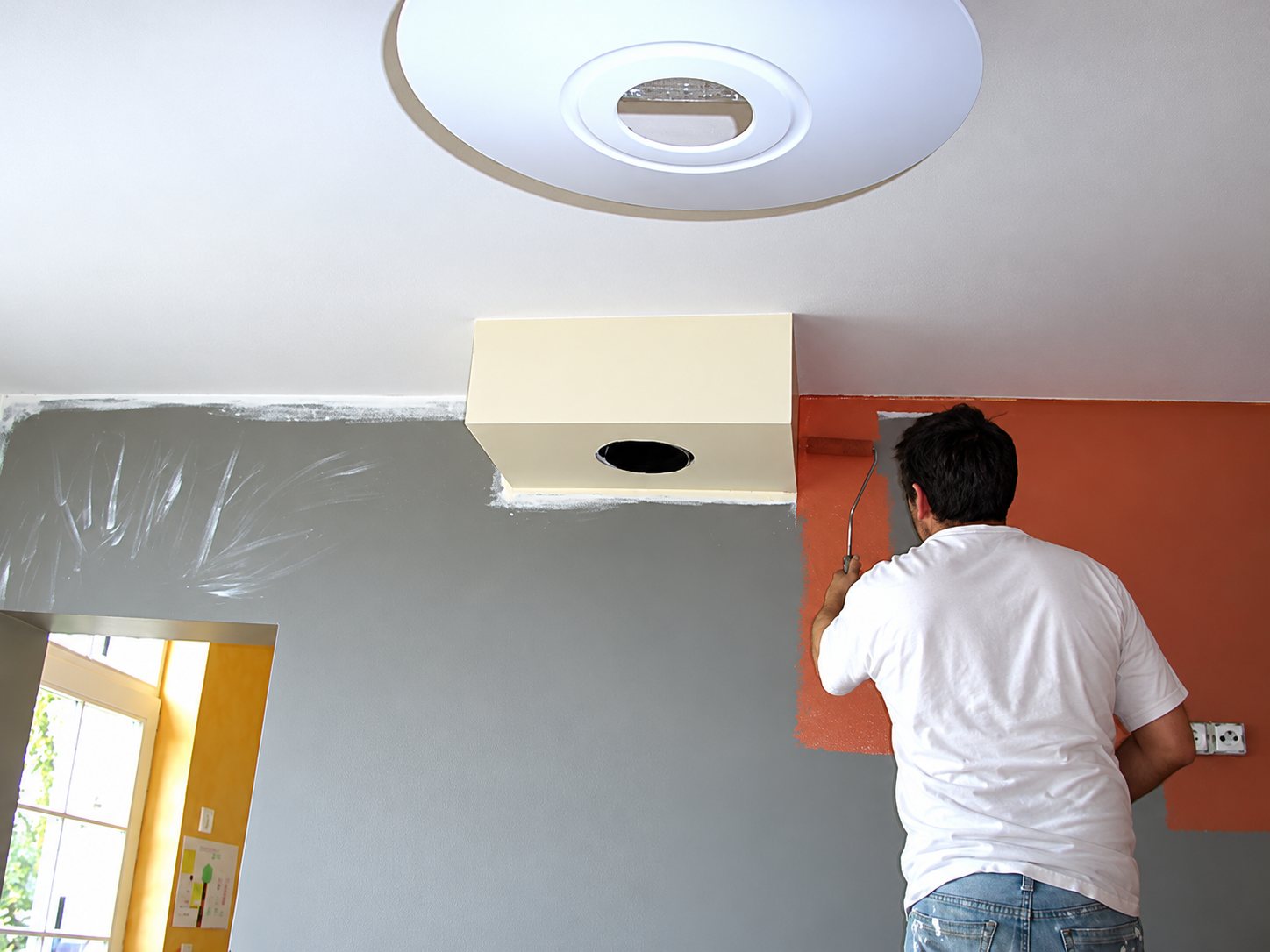 Un peintre en t-shirt blanc applique de la peinture orange sur un mur à côté d'un mur gris, sous un plafond blanc avec un luminaire rond.