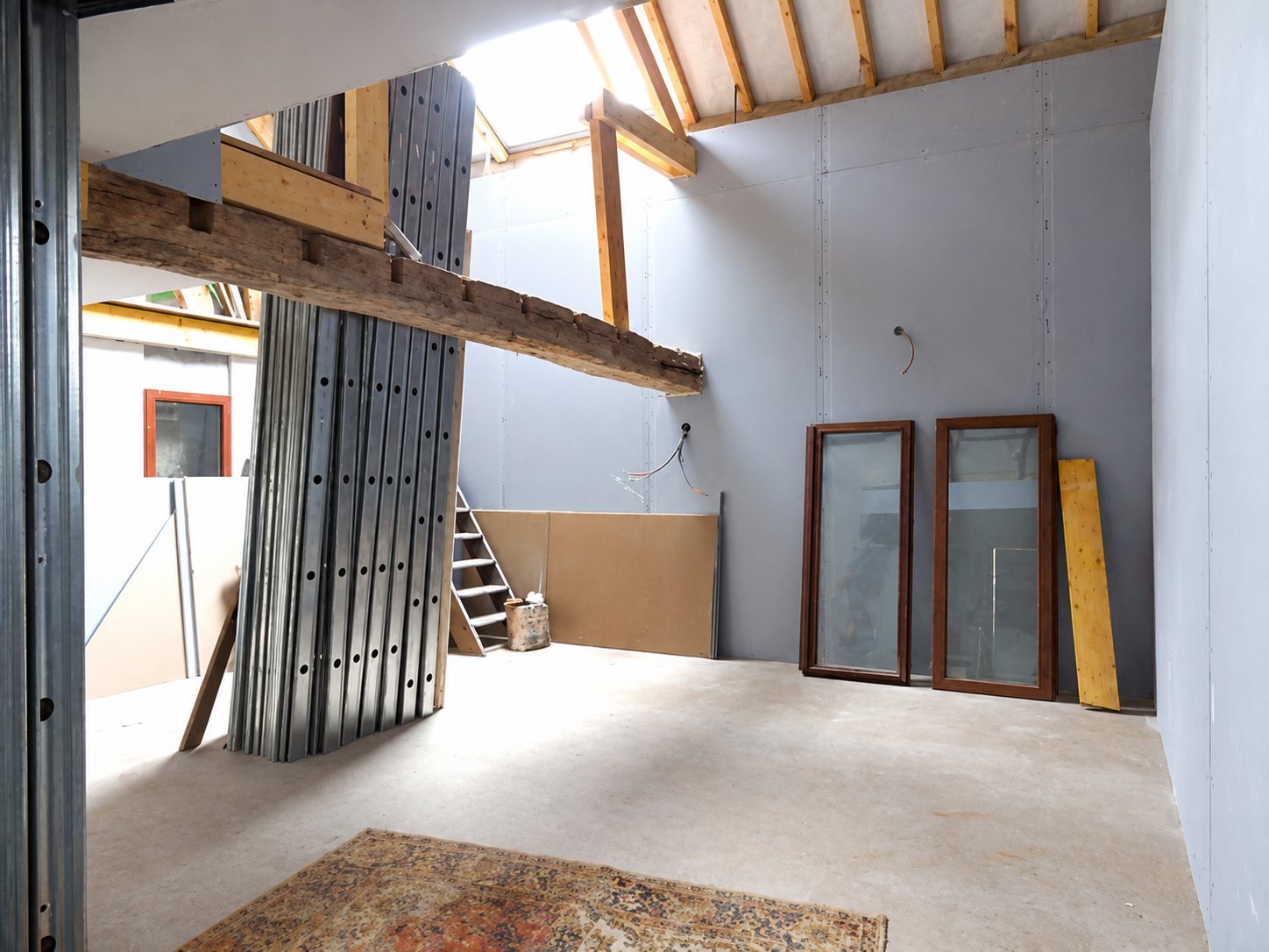 Intérieur d'une grande pièce en cours de rénovation, avec murs en plaques grises, charpente en bois apparente, deux fenêtres appuyées au mur et un tapis au sol.