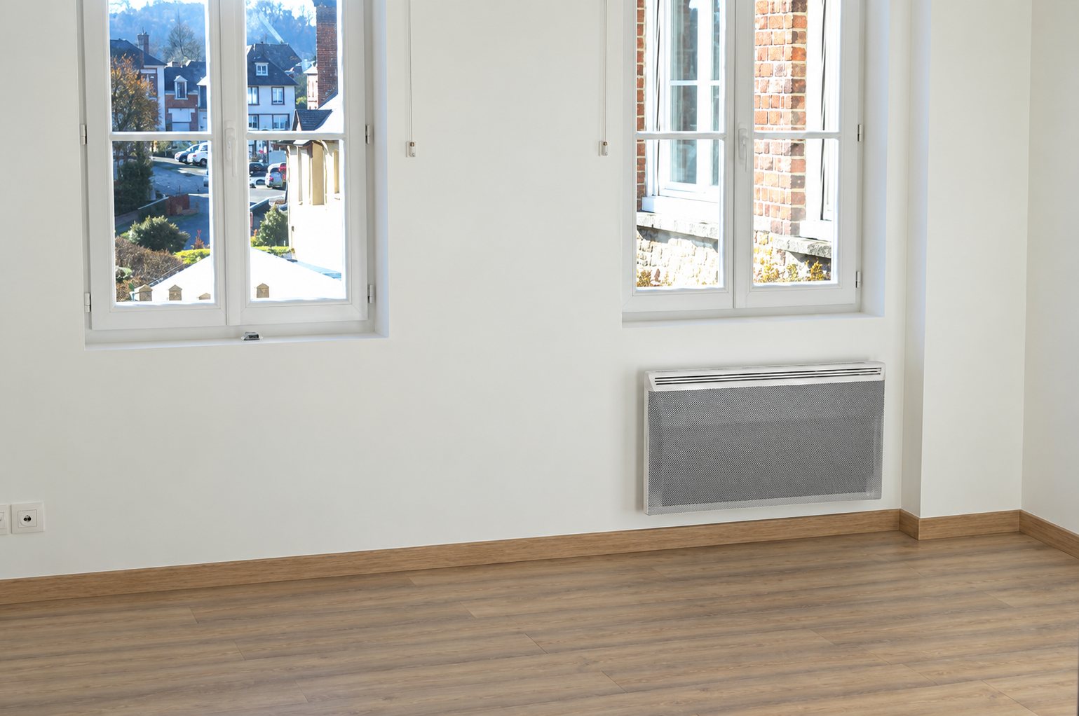 Pièce vide fraîchement rénovée aux murs blancs, avec deux fenêtres donnant sur des maisons, un radiateur électrique et un parquet stratifié.