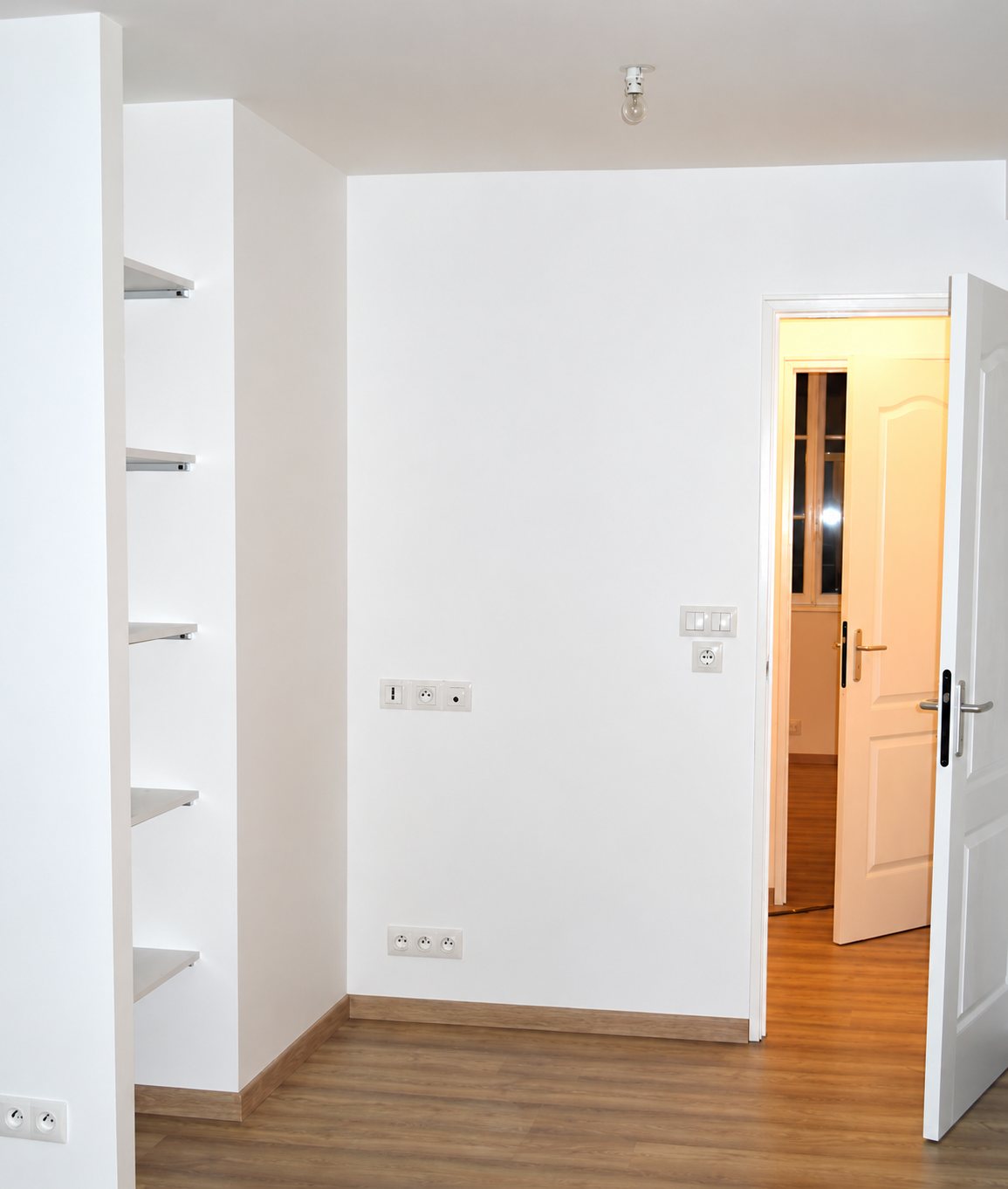 Pièce vide fraîchement rénovée aux murs blancs, avec des étagères encastrées, des prises électriques et une porte ouverte sur un couloir au parquet clair.