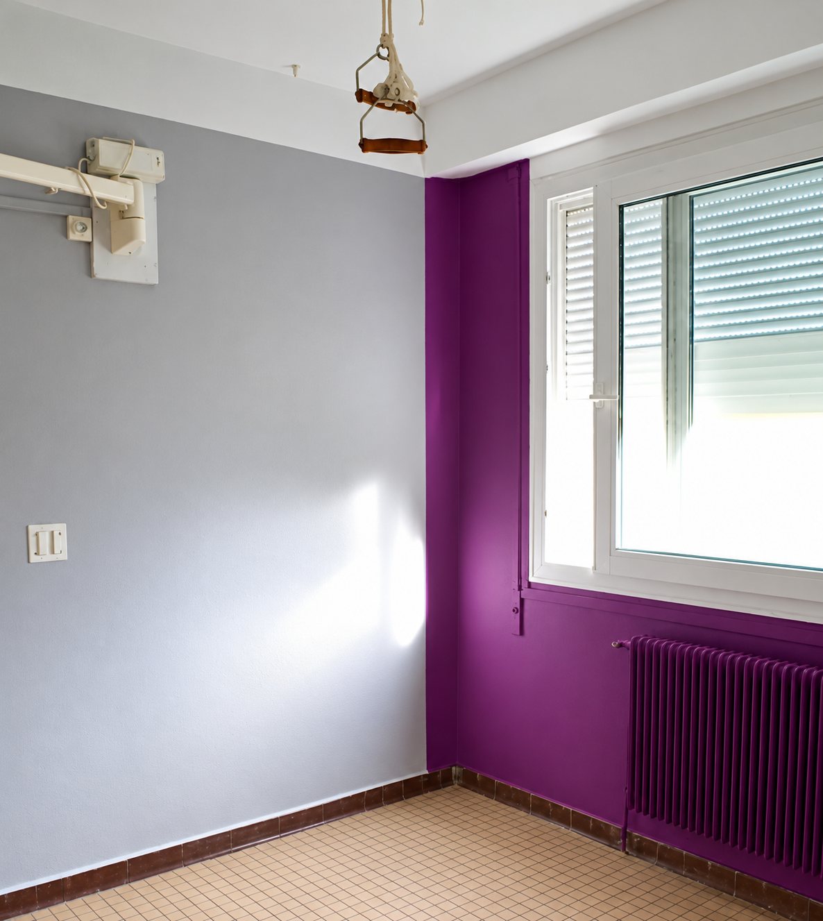 Pièce vide aux murs peints en gris clair et en violet, avec une fenêtre à volet roulant, un radiateur violet et un carrelage beige au sol.