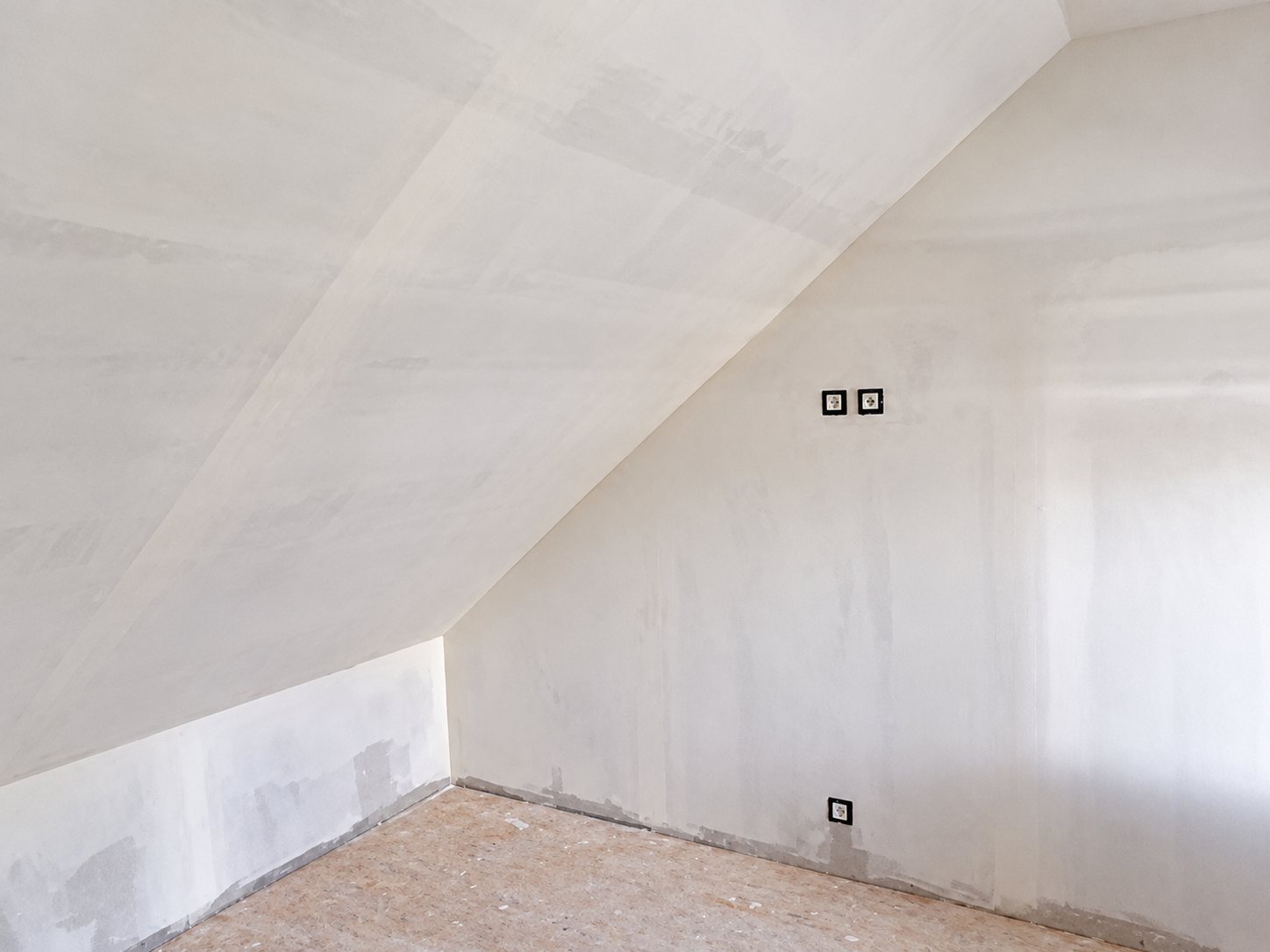 Pièce mansardée en travaux dont les murs et le plafond inclinés sont enduits de plâtre blanc, avec trois prises électriques encastrées et un sol brut.