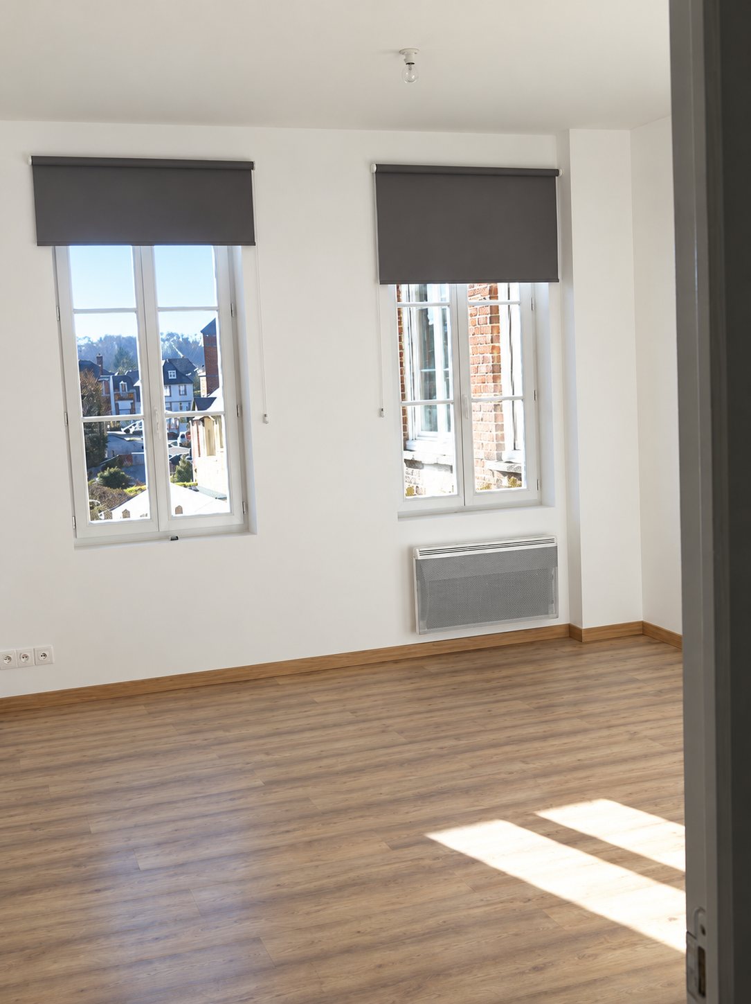 Pièce vide fraîchement rénovée aux murs blancs, avec deux fenêtres équipées de stores gris, un radiateur électrique et un parquet stratifié clair.