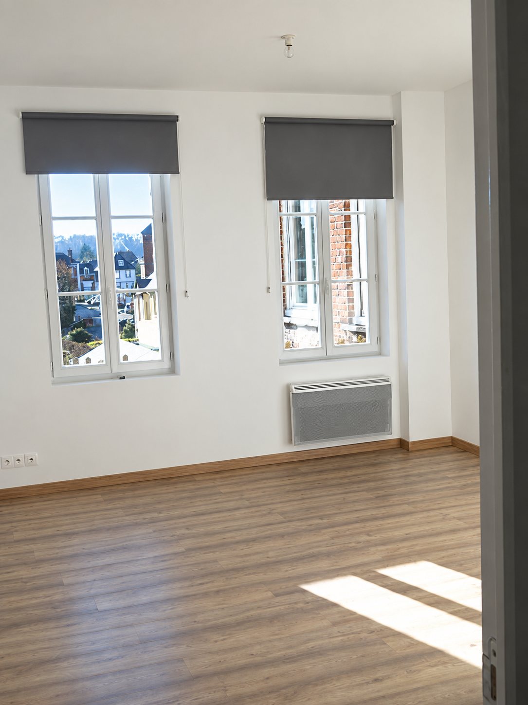 Pièce vide fraîchement rénovée aux murs blancs, avec deux fenêtres à stores gris, un radiateur électrique et un parquet stratifié clair éclairé par le soleil.