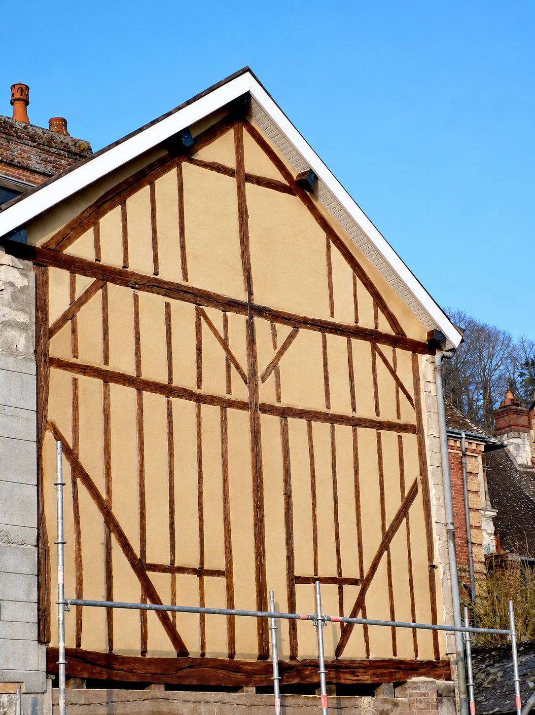 Pignon d'une maison à colombages fraîchement rénové, avec enduit beige entre les poutres en bois brun, un rebord de toit blanc et un échafaudage au premier plan.
