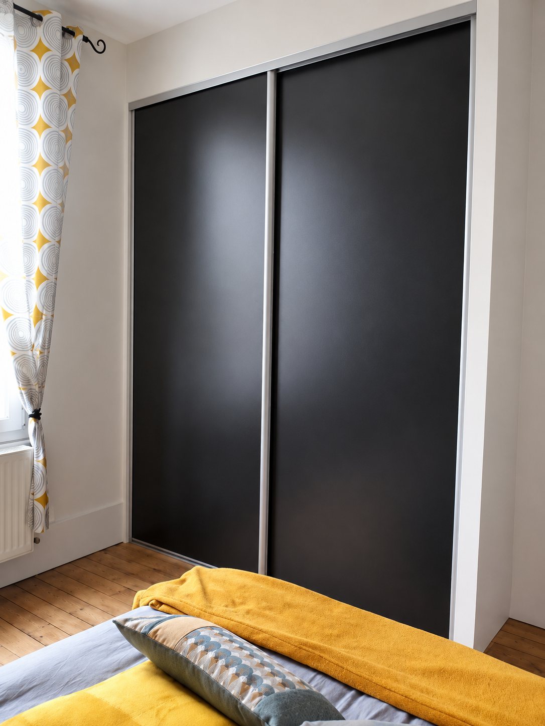 Dans une chambre au parquet ancien, un placard encastré à deux portes coulissantes noires est installé près d'une fenêtre ornée d'un rideau à motifs jaunes, avec un lit au premier plan.