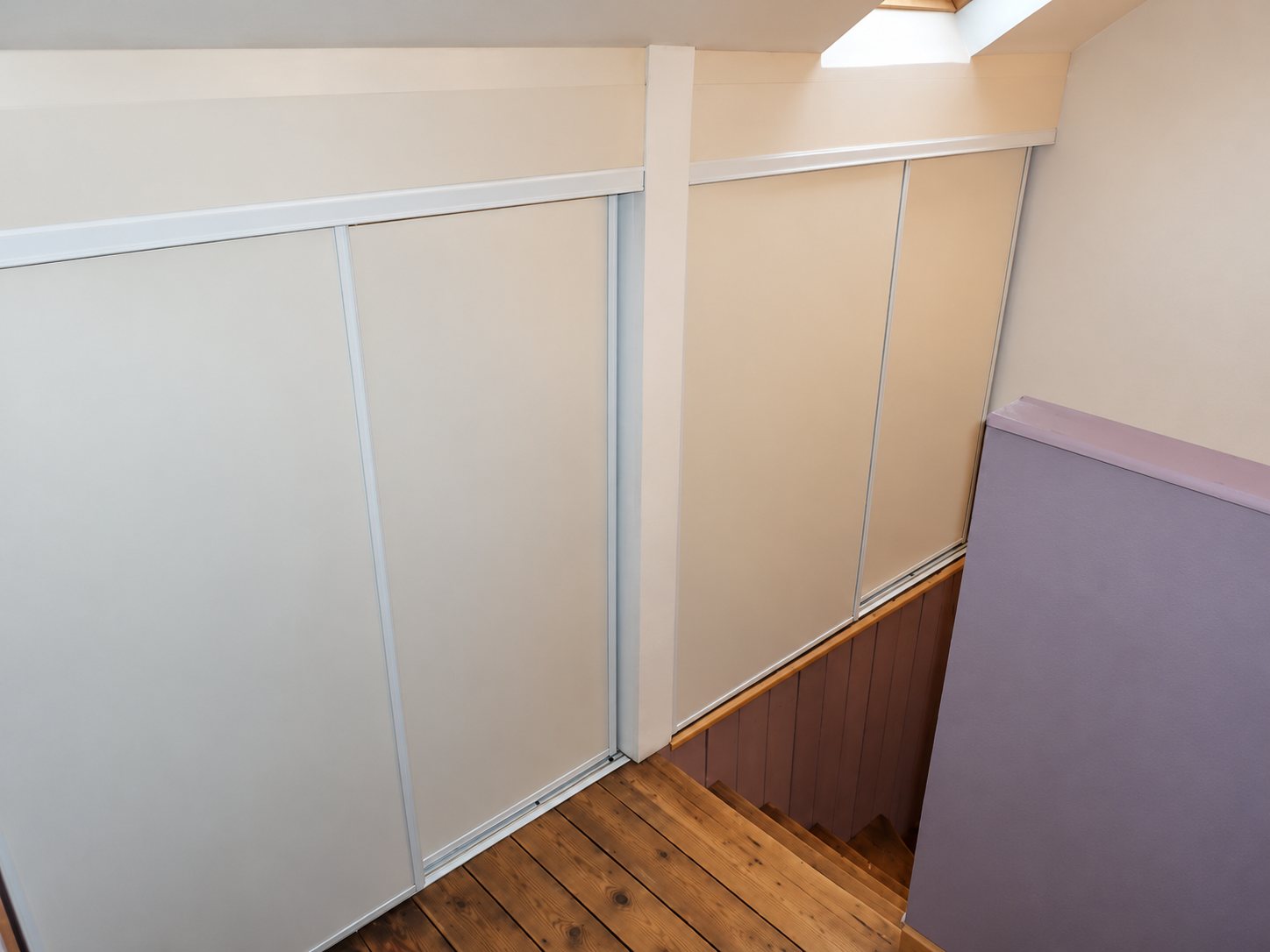 Des portes de placard coulissantes blanches sont installées sous un plafond en pente, à côté d'un escalier au plancher de bois et d'un mur violet.