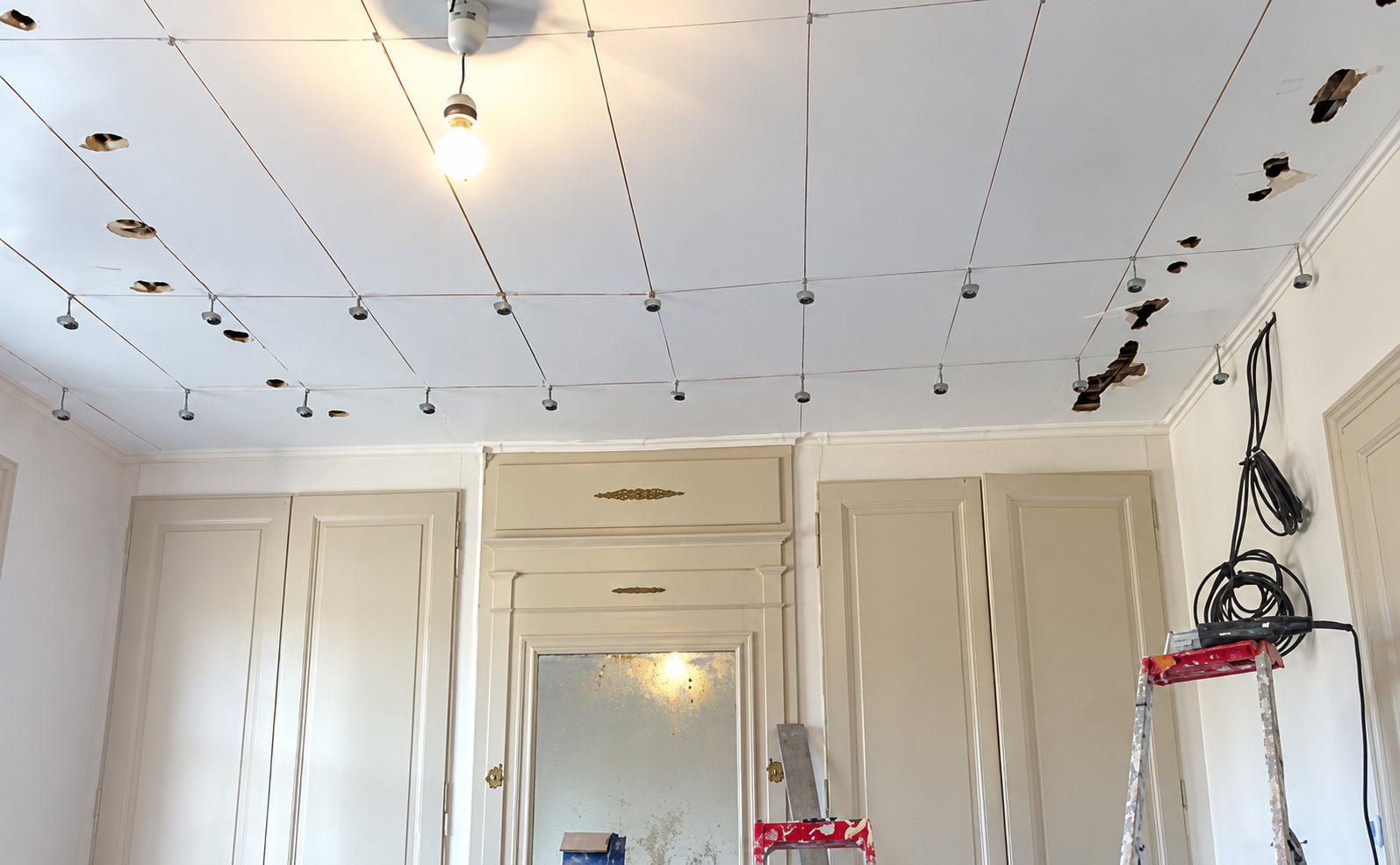 Une pièce en rénovation avec un plafond blanc percé de petits trous et de fils, des placards anciens au fond et une échelle rouge au premier plan.