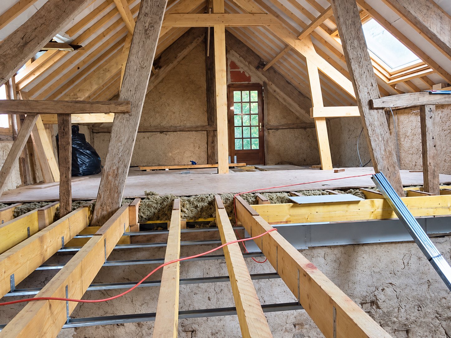 Combles en cours de rénovation avec charpente en bois, solives neuves, isolation en vrac et fenêtre de toit.