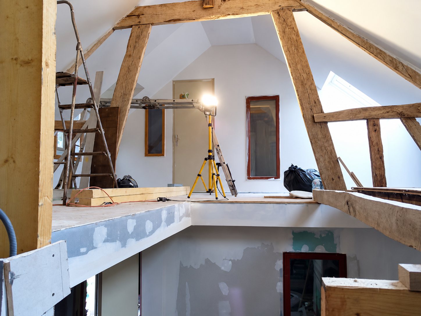 Intérieur de combles en cours de rénovation avec poutres en bois apparentes, murs blancs, échelle et projecteur de chantier jaune sur trépied.