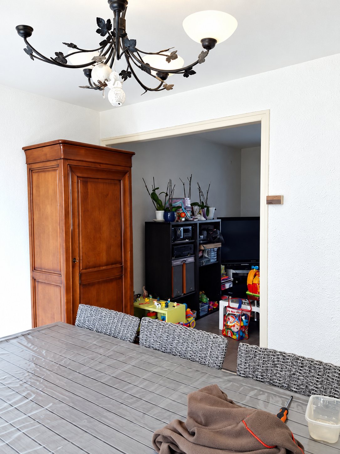 Une salle à manger avec une table grise, des chaises en rotin tressé, une armoire en bois et un lustre en fer forgé, ouverte sur un salon avec des jouets d'enfant.