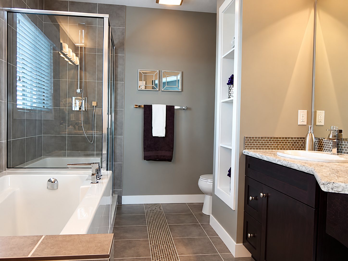 Salle de bain aux murs gris avec une baignoire blanche, une douche vitrée, des serviettes suspendues et un meuble vasque brun foncé avec plan en granit.