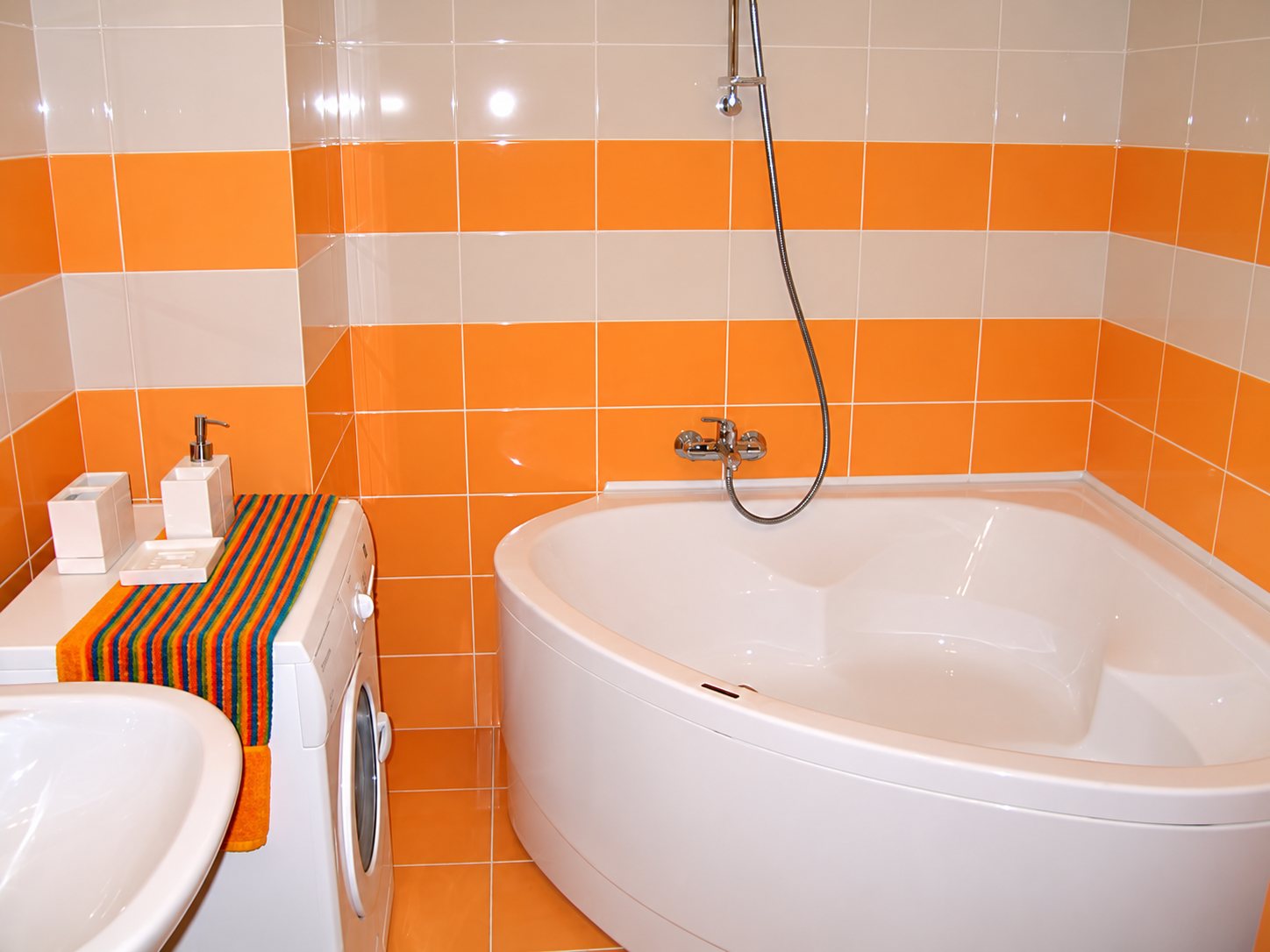 Salle de bain aux murs carrelés orange et blanc avec une grande baignoire d'angle, un lave-linge recouvert d'un tapis rayé et un lavabo au premier plan.