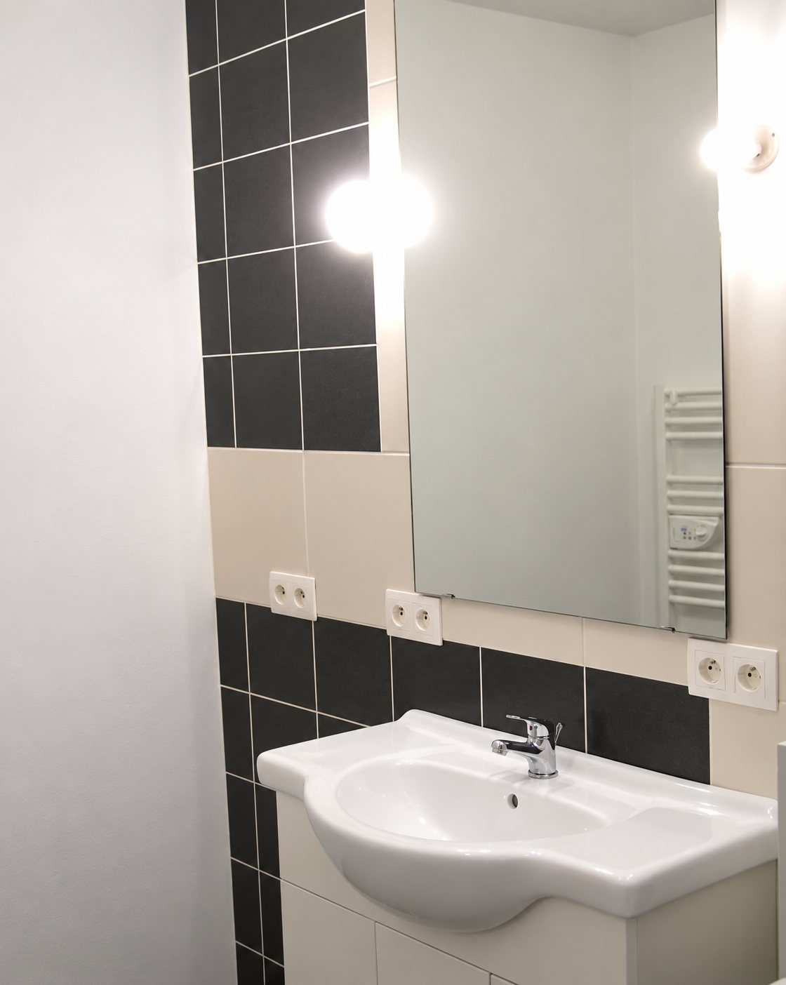 Salle de bain rénovée avec meuble vasque blanc, grand miroir rectangulaire, sèche-serviettes et carrelage gris foncé et beige.