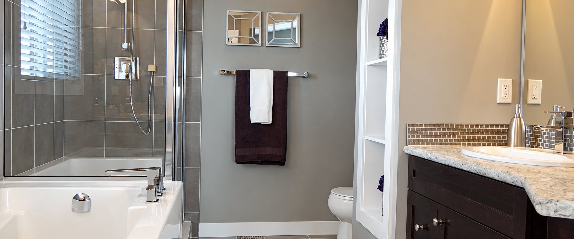 Salle de bains moderne aux murs gris avec une douche vitrée, une baignoire blanche, des serviettes suspendues et un meuble vasque en bois foncé avec plan de travail en granit.