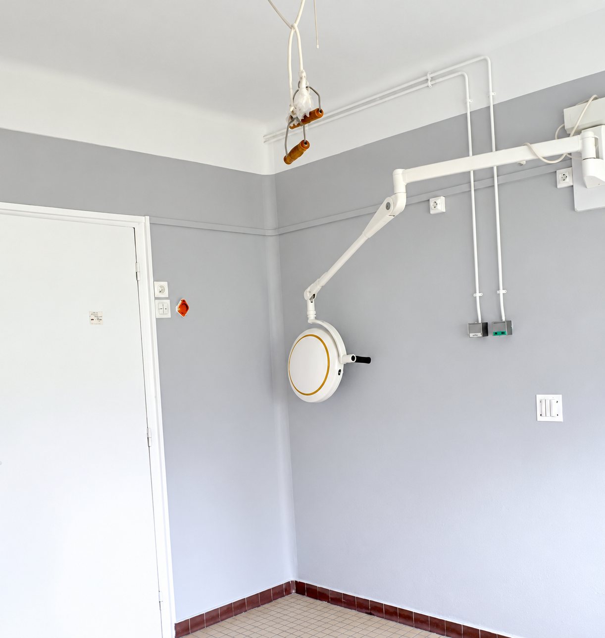 Une pièce vide aux murs gris clair et à la porte blanche, équipée d'une lampe d'examen articulée fixée au mur et de câbles apparents.