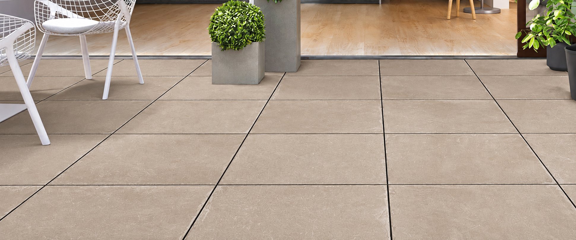 Une terrasse recouverte de grandes dalles de carrelage beige, décorée de chaises blanches ajourées et de plantes en pots, devant une grande baie vitrée.